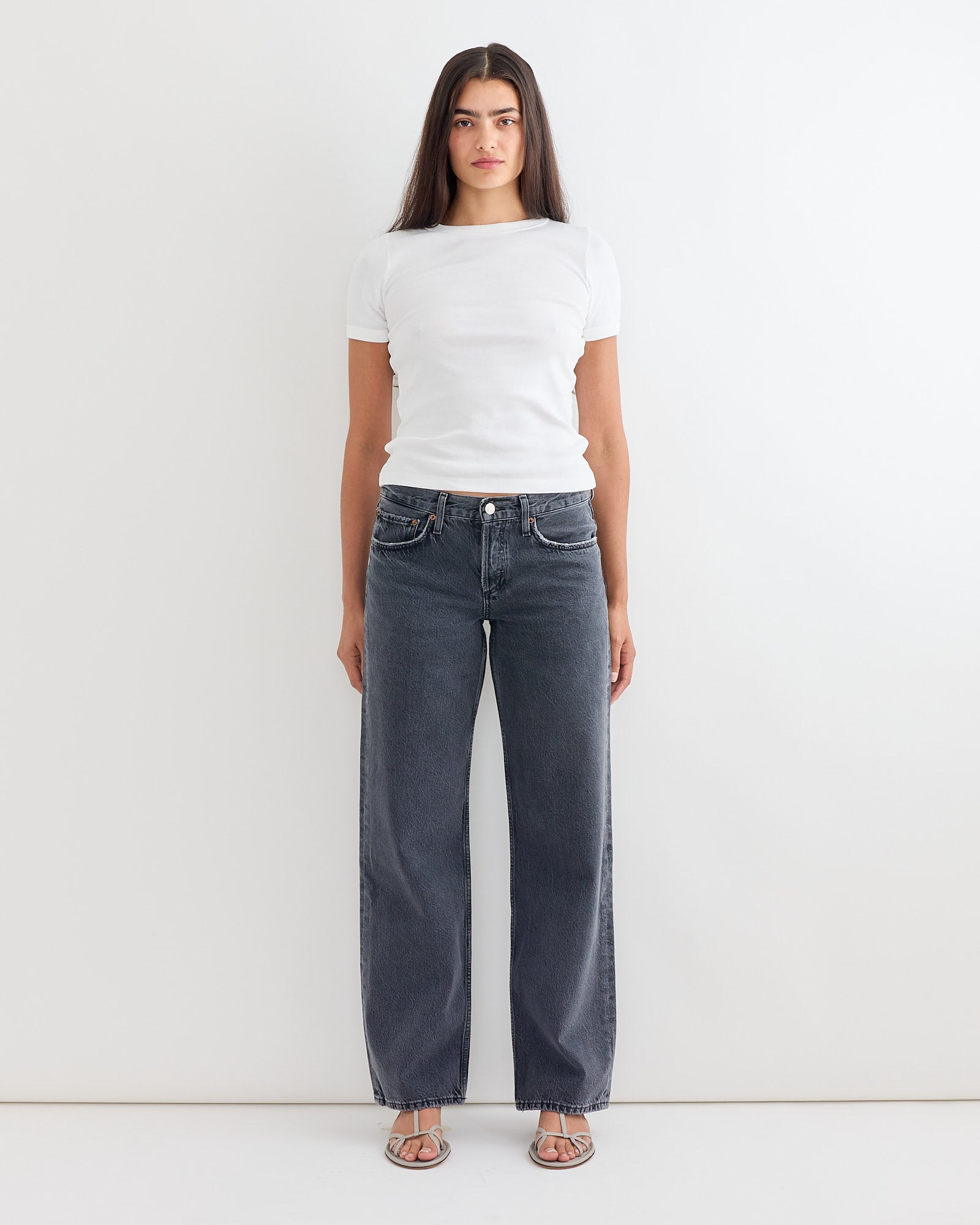 A woman stands against a white wall wearing AGOLDE’s Low Rise Loose Jean in Symbolic, paired with a fitted white tee and sandals. Her relaxed pose and straight hair highlight the comfortable, easy fit of her outfit.