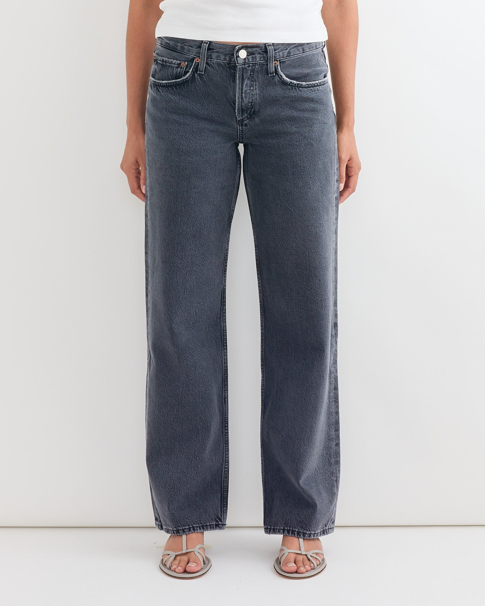 A person models the AGOLDE Low Rise Loose Jean in Symbolic with a white top and open-toed sandals against a plain background—ideal for viewing sizing and fit details before your next online chat or purchase.