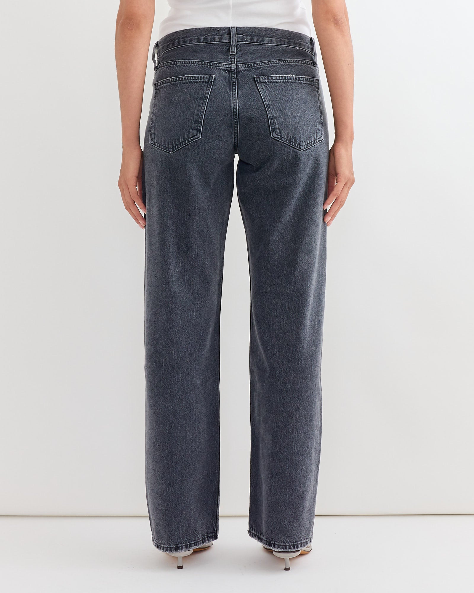 A person wearing AGOLDEs Low Rise Loose Jean in Symbolic, paired with a white top, stands facing a white wall with their back to the camera.
