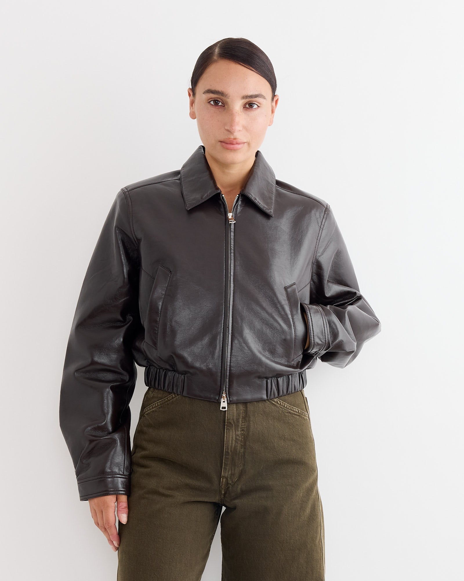 A person with dark hair pulled back wears the AGOLDE Essie Recycled Cropped Jacket in Chocolate and olive green pants, standing against a plain white background with a neutral expression.