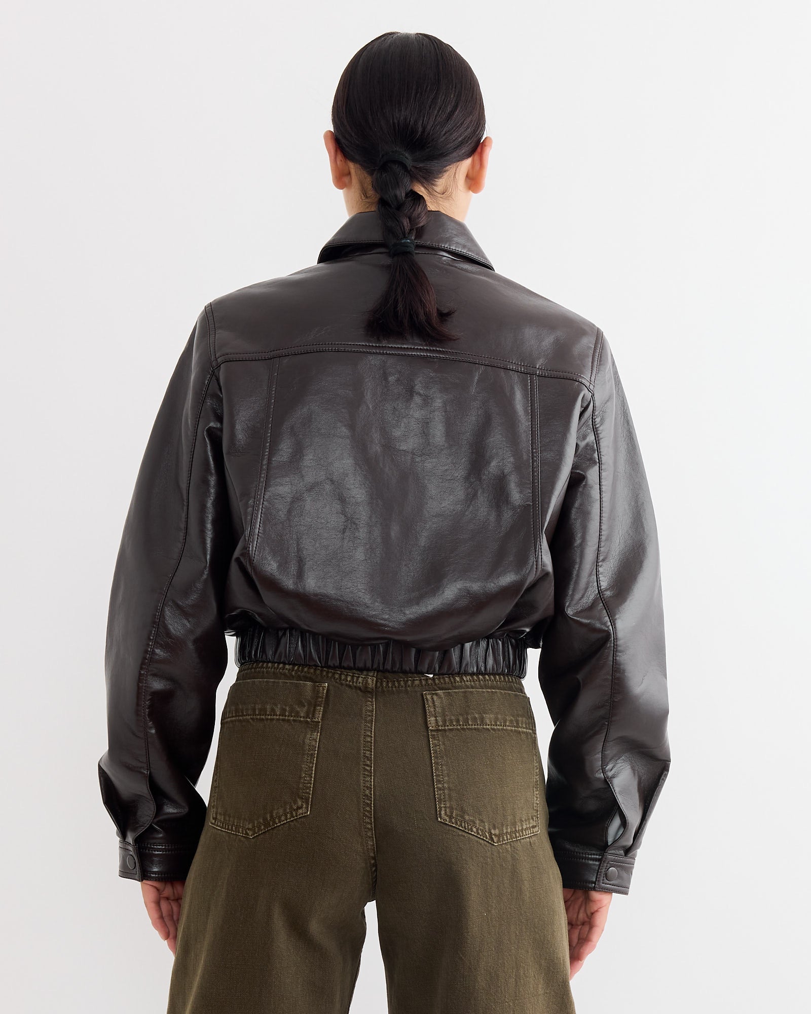 A person with dark hair in a low braid is seen from behind, wearing the Essie Recycled Cropped Jacket in Chocolate by AGOLDE and olive green high-waisted pants against a plain white background.