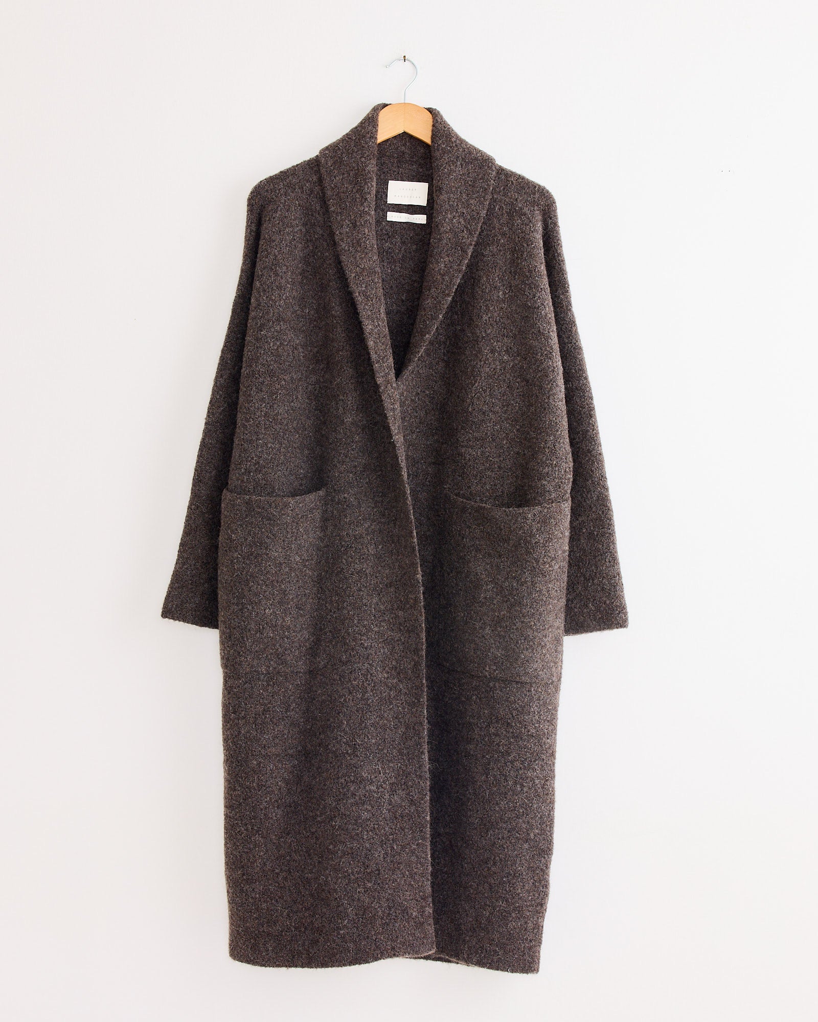 The Lauren Manoogian Double Face Long Coat in Blackened, featuring a shawl collar and two large front pockets, hangs on a wooden hanger against a white wall.