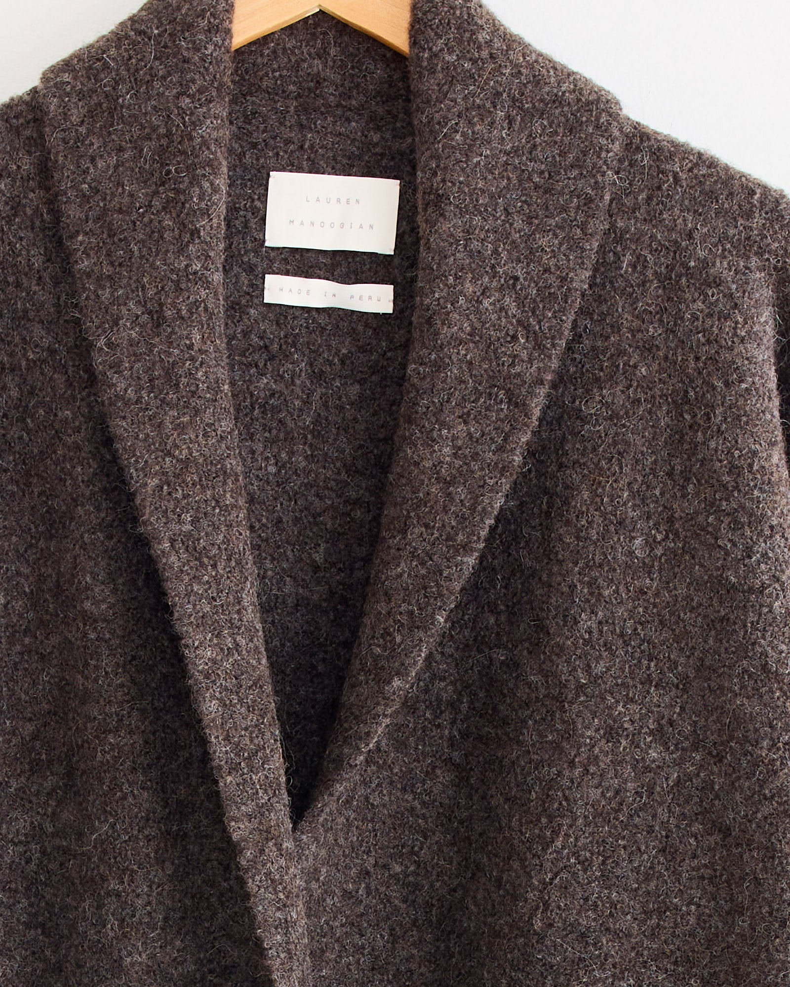 A close-up of the Lauren Manoogian Double Face Long Coat in Blackened hanging on a wooden hanger, highlighting its textured fabric, collar, and an inside white label.