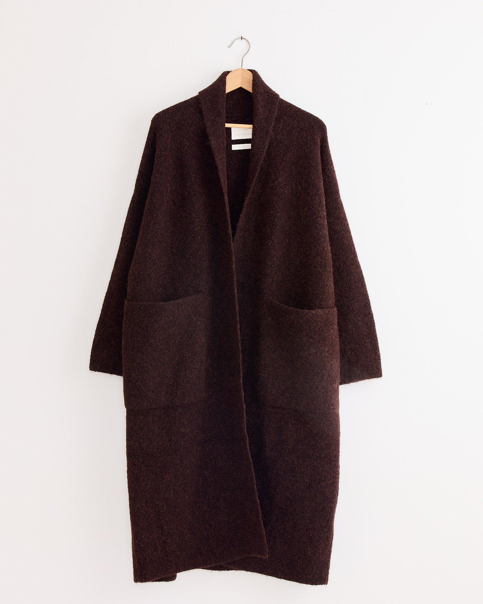 A Lauren Manoogian Double Face Long Coat in Merlot hangs on a wooden hanger against a plain white wall, featuring large front pockets.