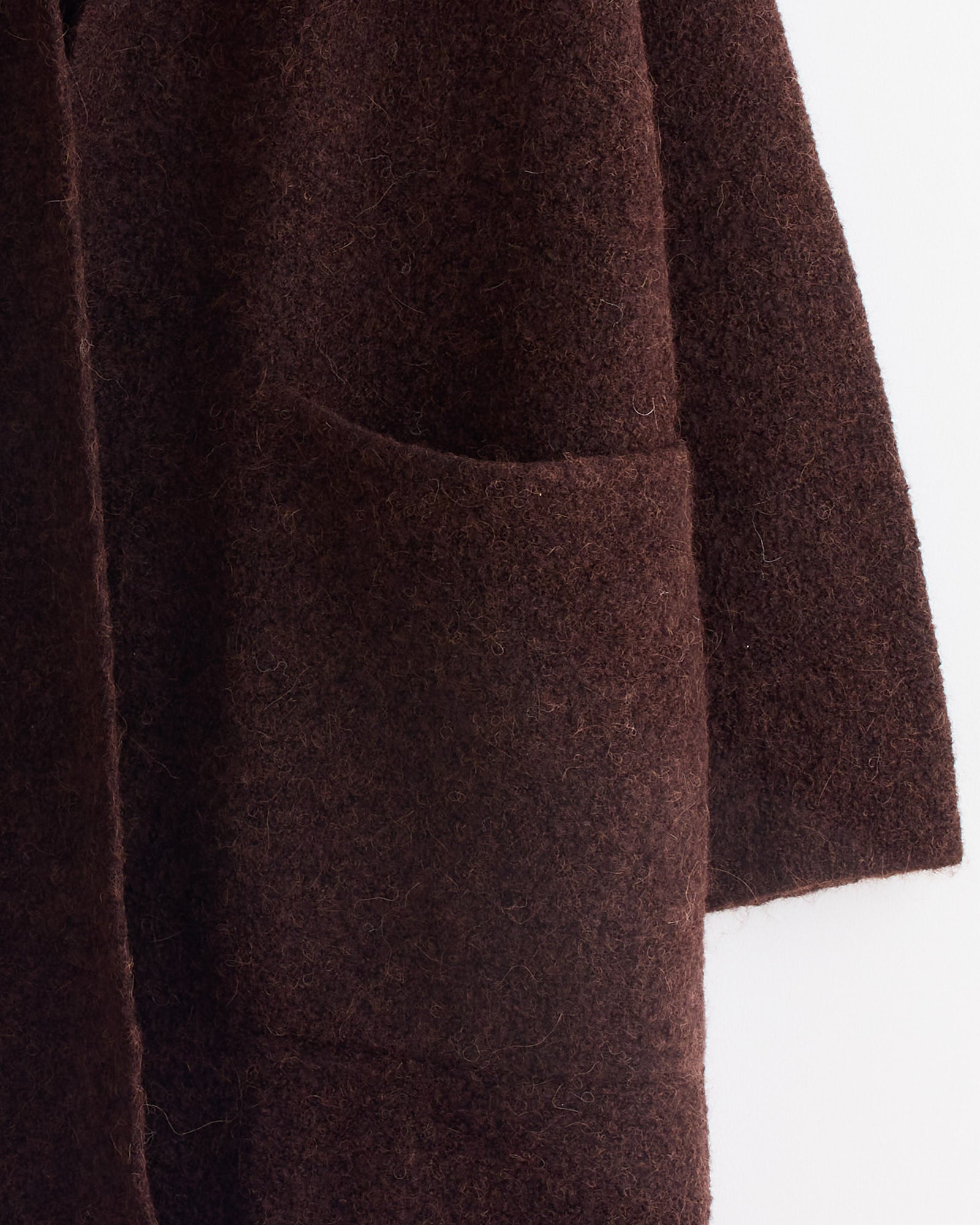 A close-up of the Lauren Manoogian Double Face Long Coat in Merlot, showing its textured dark fabric, large front pocket, and visible sleeve, set against a light background.