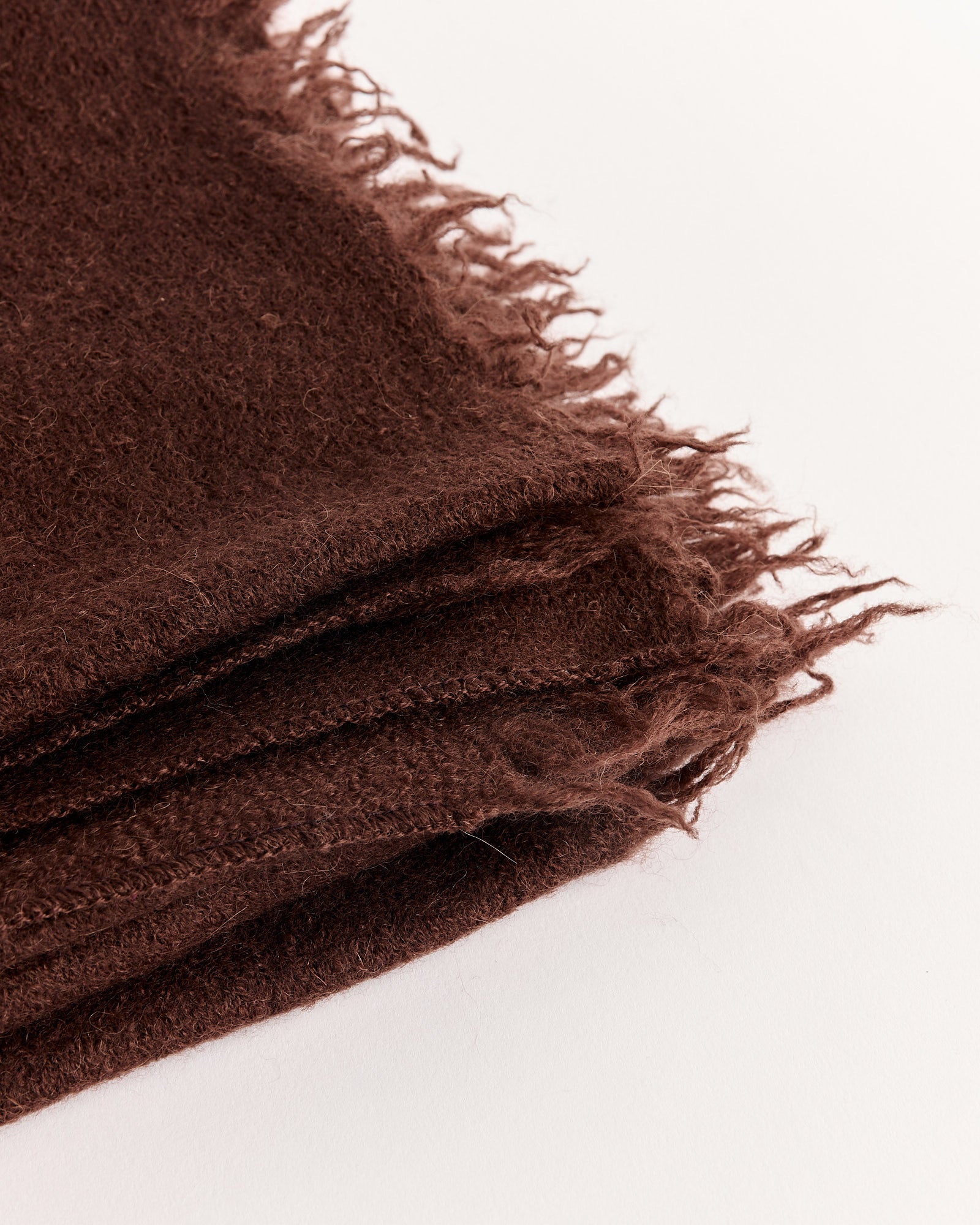 A close-up of the Lauren Manoogian Felt Fringe Scarf in dark brown, featuring soft felted wool with frayed edges, folded and placed on a white background.