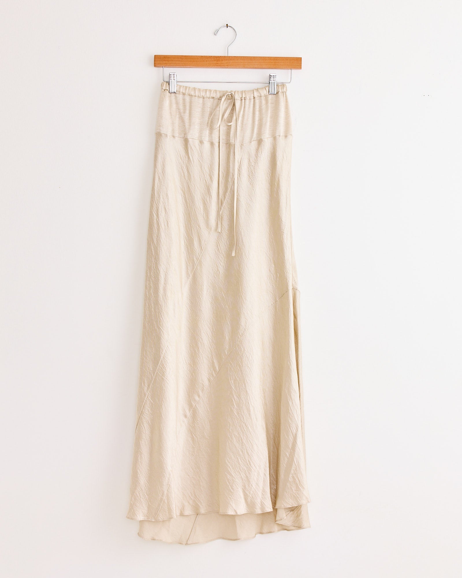 The Lauren Manoogian New Luster Skirt in Oyster is a long, beige, high-waisted skirt with a drawstring waistband and subtle crinkle texture. Made in Japan, it features a slightly flared hem and hangs on a wooden hanger against a white wall.