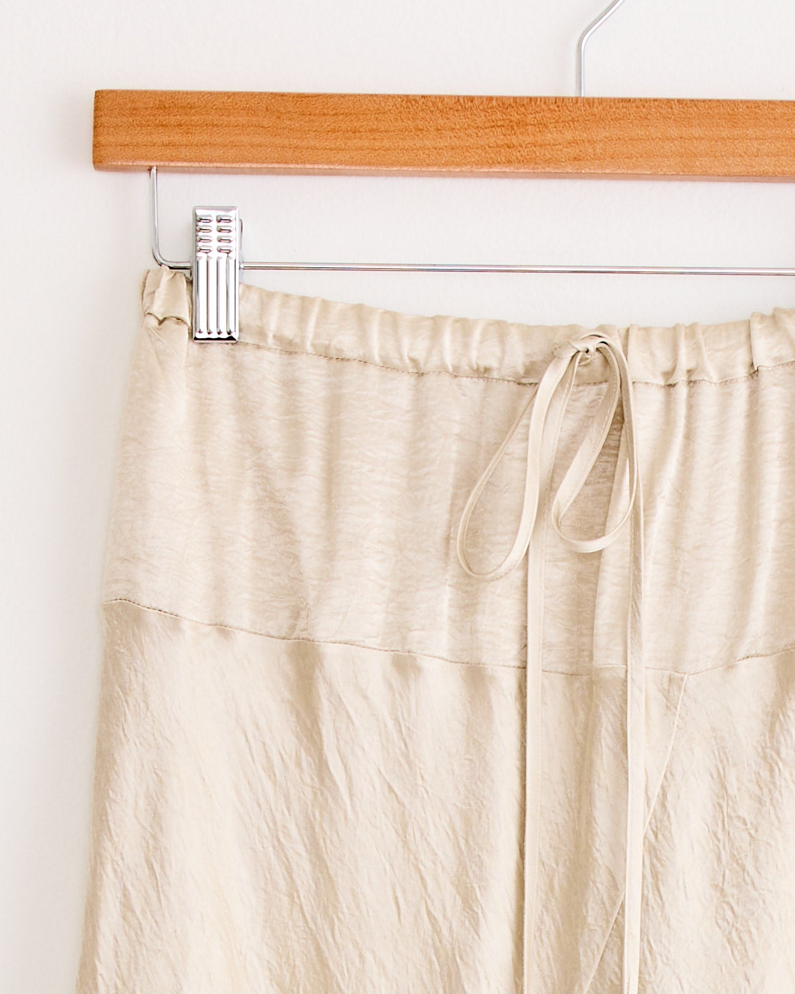 A close-up of the Lauren Manoogian New Luster Skirt in Oyster, featuring a crinkled texture, elastic waistband, and drawstring, shown on a wooden hanger against a white wall.