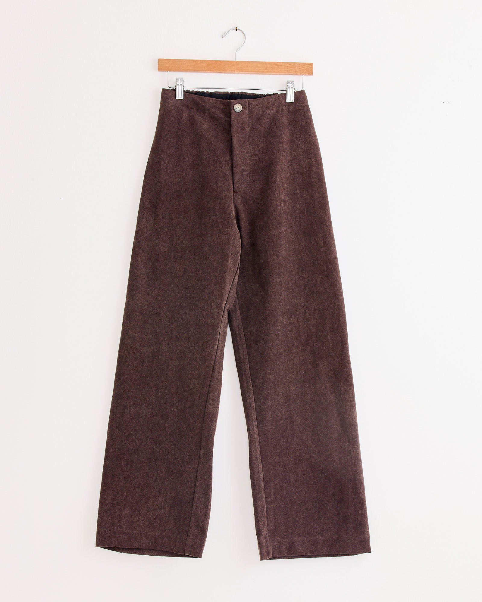 A pair of Lauren Manoogian Canvas Gallery Pant in Washed Soil, featuring a high-waisted, wide-leg design, is displayed on a wooden hanger against a plain white wall.