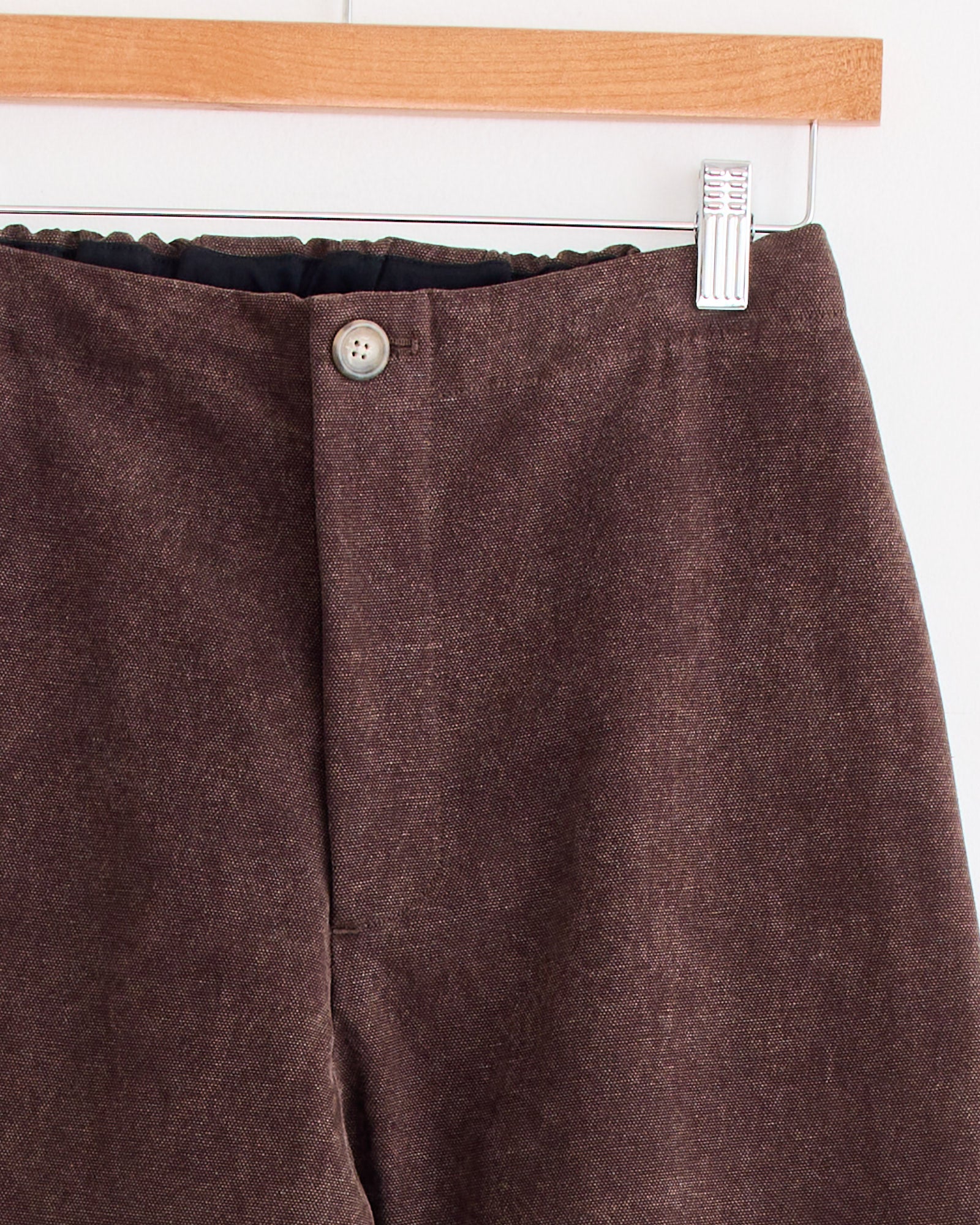 Canvas Gallery Pant in Washed Soil by Lauren Manoogian hangs on a wooden hanger, partially visible from the waist down, featuring a single button and zipper fly closure.