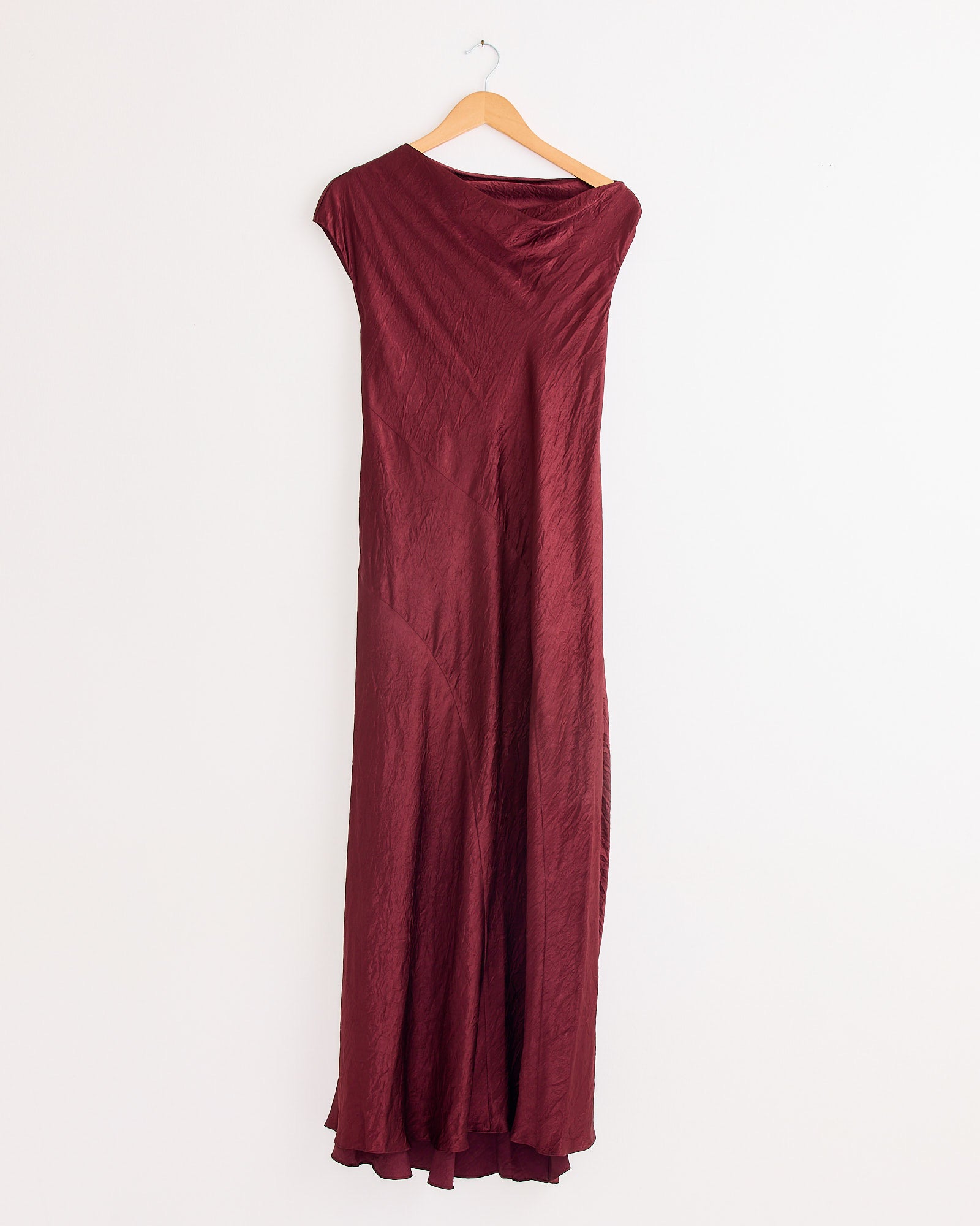 The New Luster Dress in Merlot by Lauren Manoogian, a long, sleeveless piece with a subtle sheen and draped bias fit, hangs on a wooden hanger against a plain white wall.