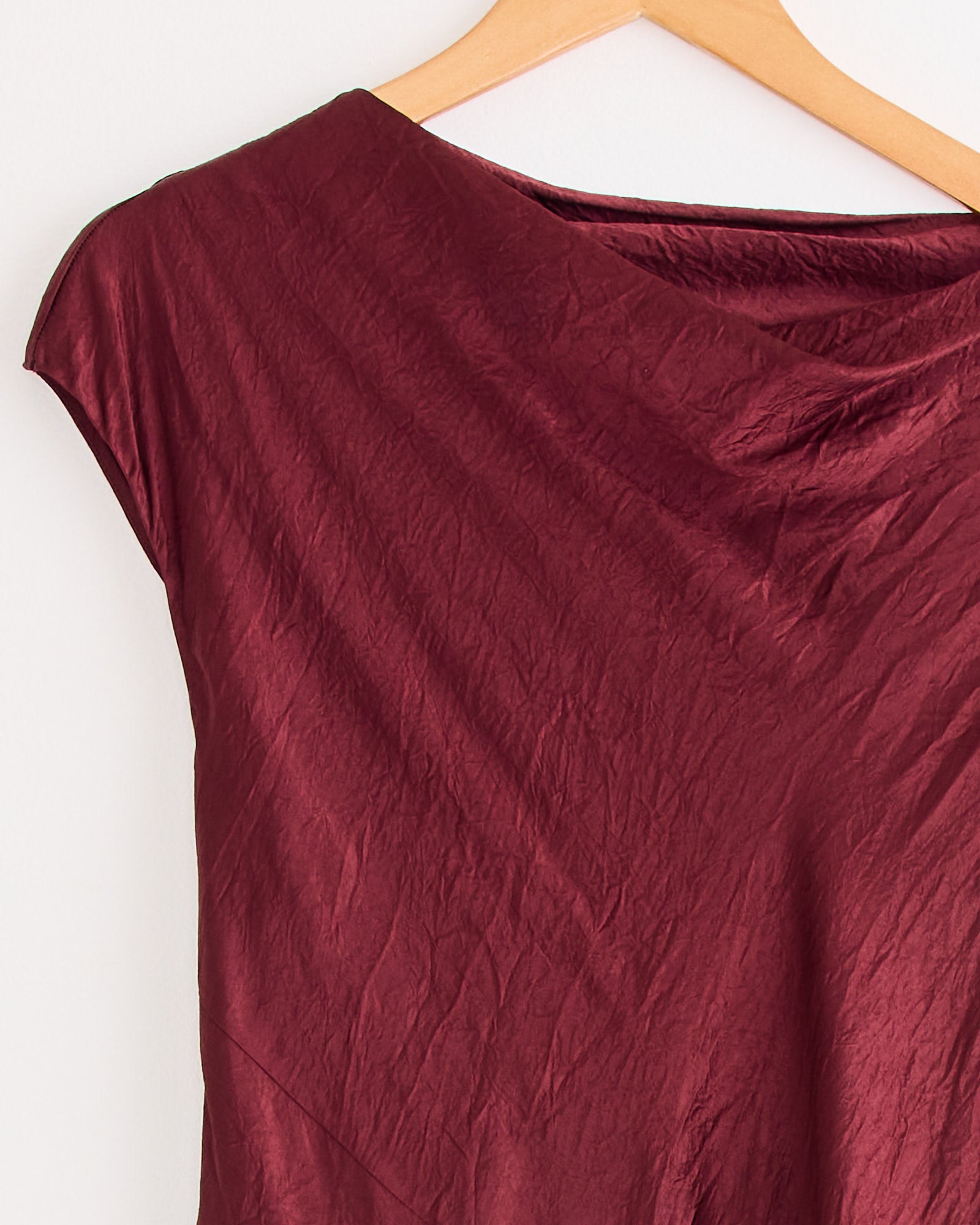 A close-up of the New Luster Dress in Merlot by Lauren Manoogian, featuring its draped bias fit and smooth finish, hanging on a wooden hanger against a white background.