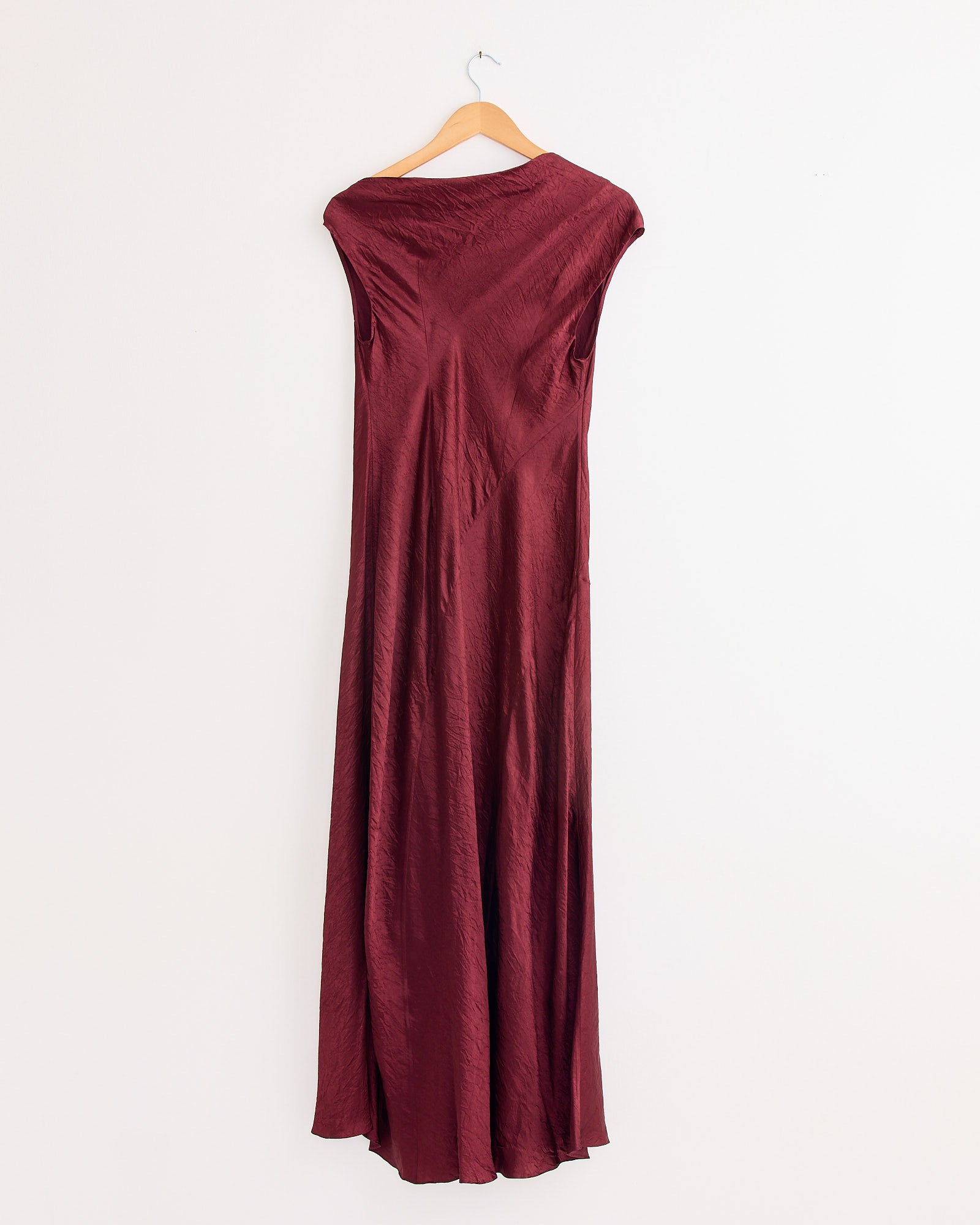 The New Luster Dress in Merlot by Lauren Manoogian, a long sleeveless asymmetrical dress with shiny textured fabric and a draped bias fit, hangs on a wooden hanger against a plain white wall.