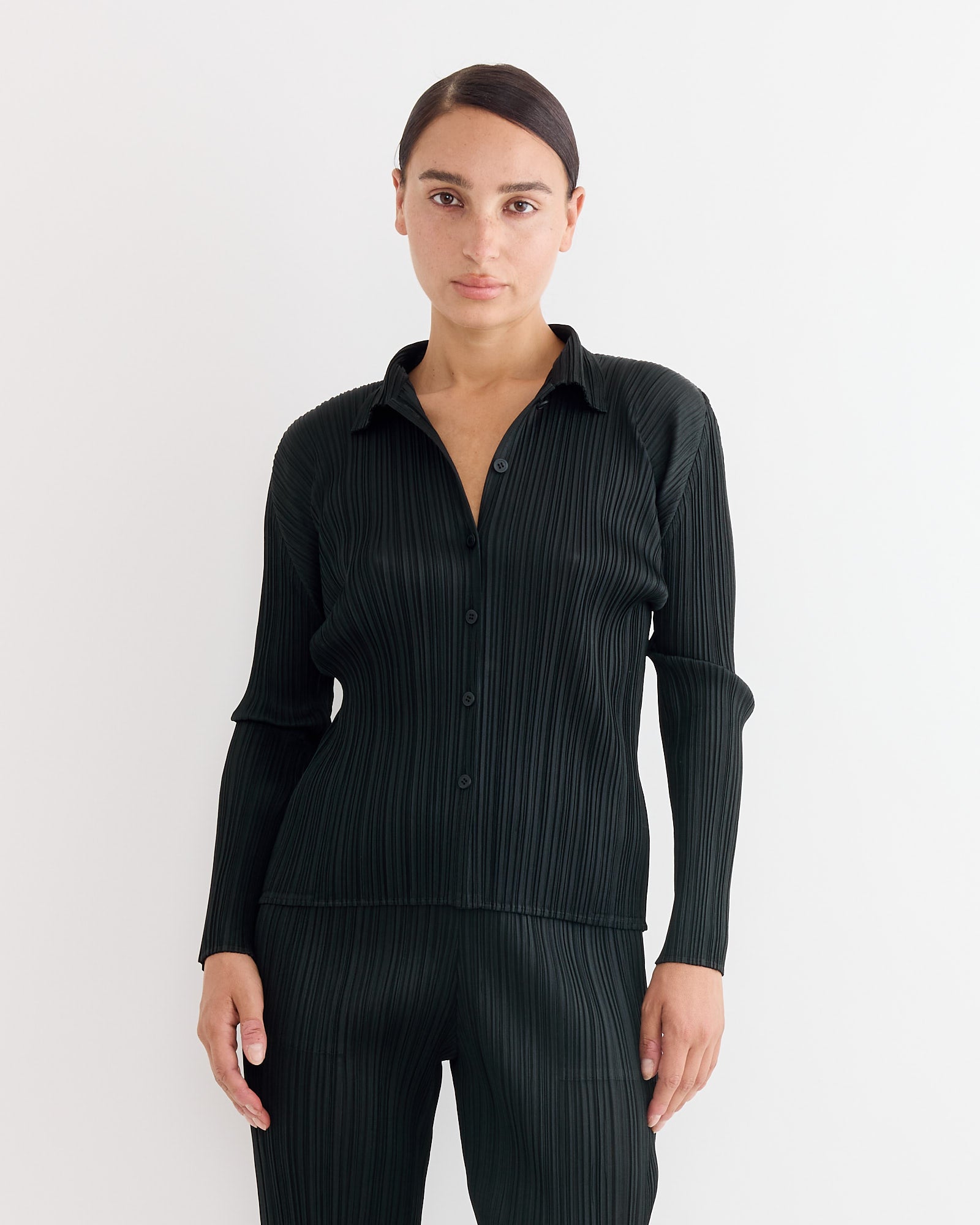 Against a plain white background, a person with dark hair pulled back wears the Basics Shirt in Black by Pleats Please Issey Miyake.