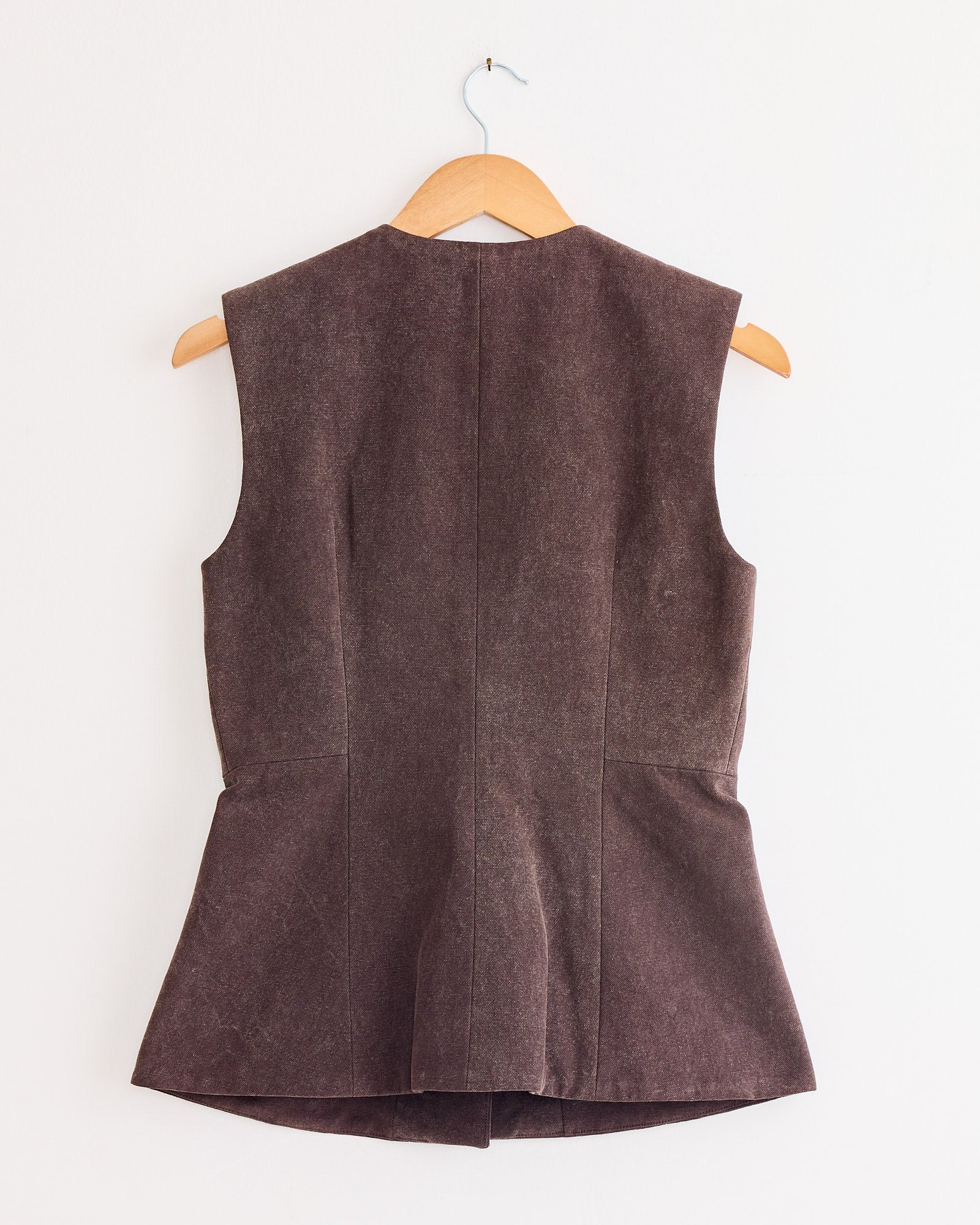 The Lauren Manoogian Canvas Bodice in Washed Soil—a sleeveless, brown tailored vest with a structured silhouette—is shown on a wooden hanger against a white wall, displayed from the back to highlight its subtle seams and flared hem.