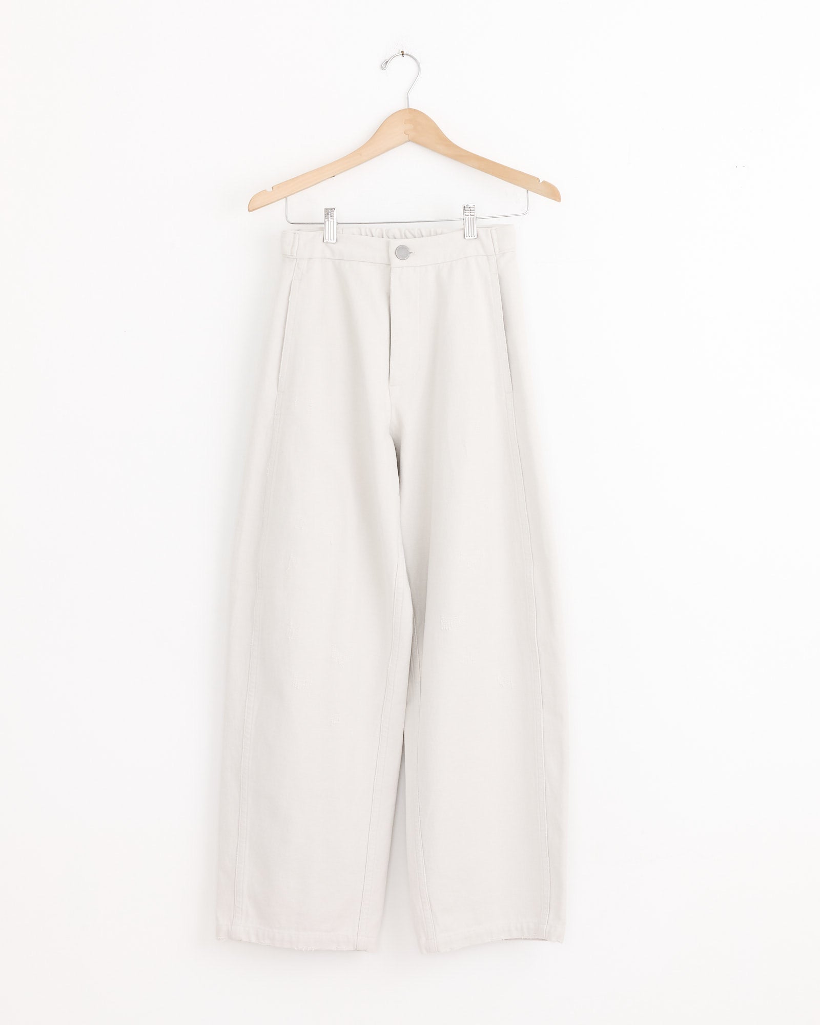 A pair of Lauren Manoogian Worn Pant in Worn Chalk, featuring a high waist and relaxed full-leg silhouette, hangs on a wooden hanger against a plain white background.
