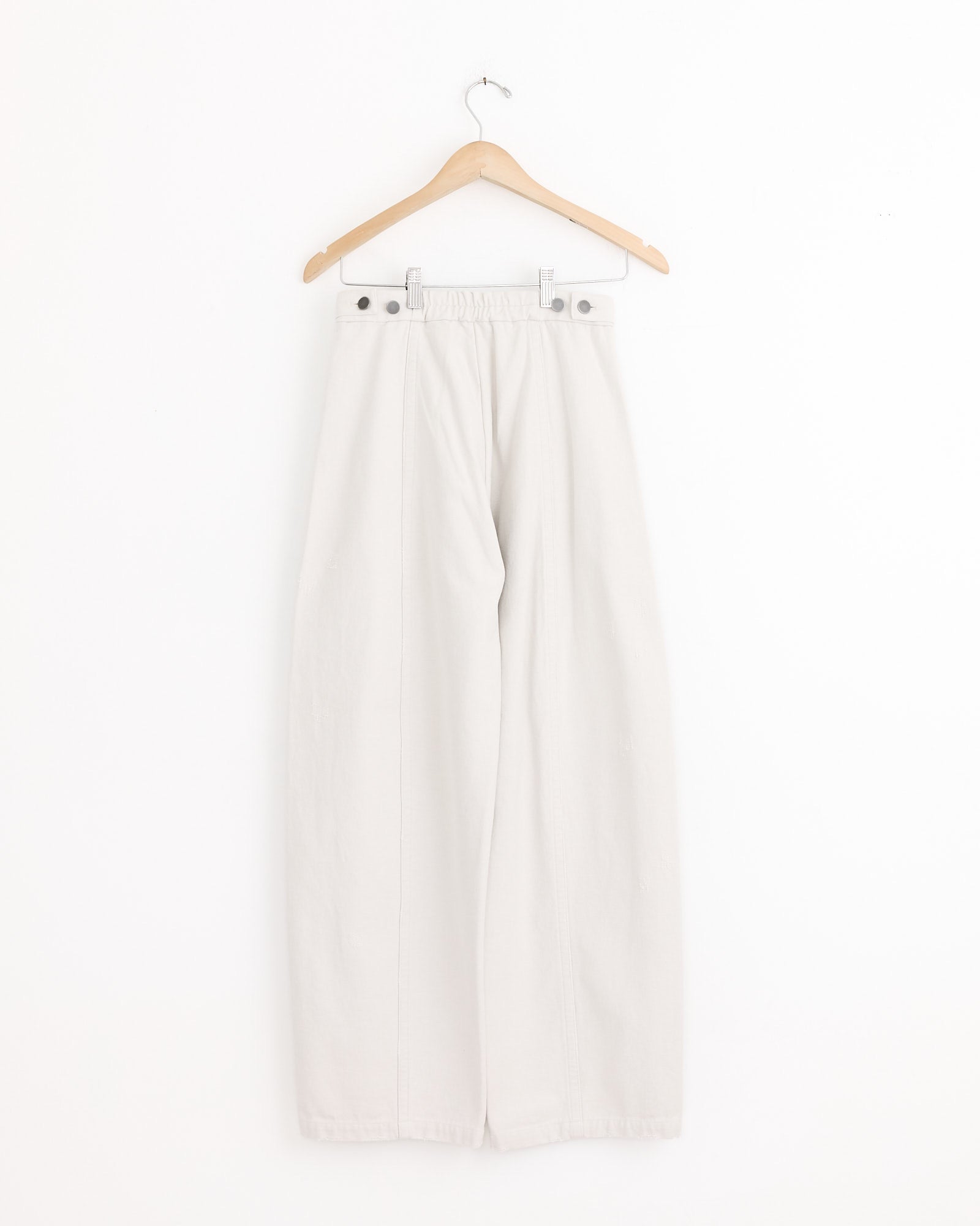 The Lauren Manoogian Worn Pant in Worn Chalk is a wide-leg, high-waisted 100% cotton style with button details at the waistband and a relaxed full-leg silhouette, shown on a wooden hanger against a white background.