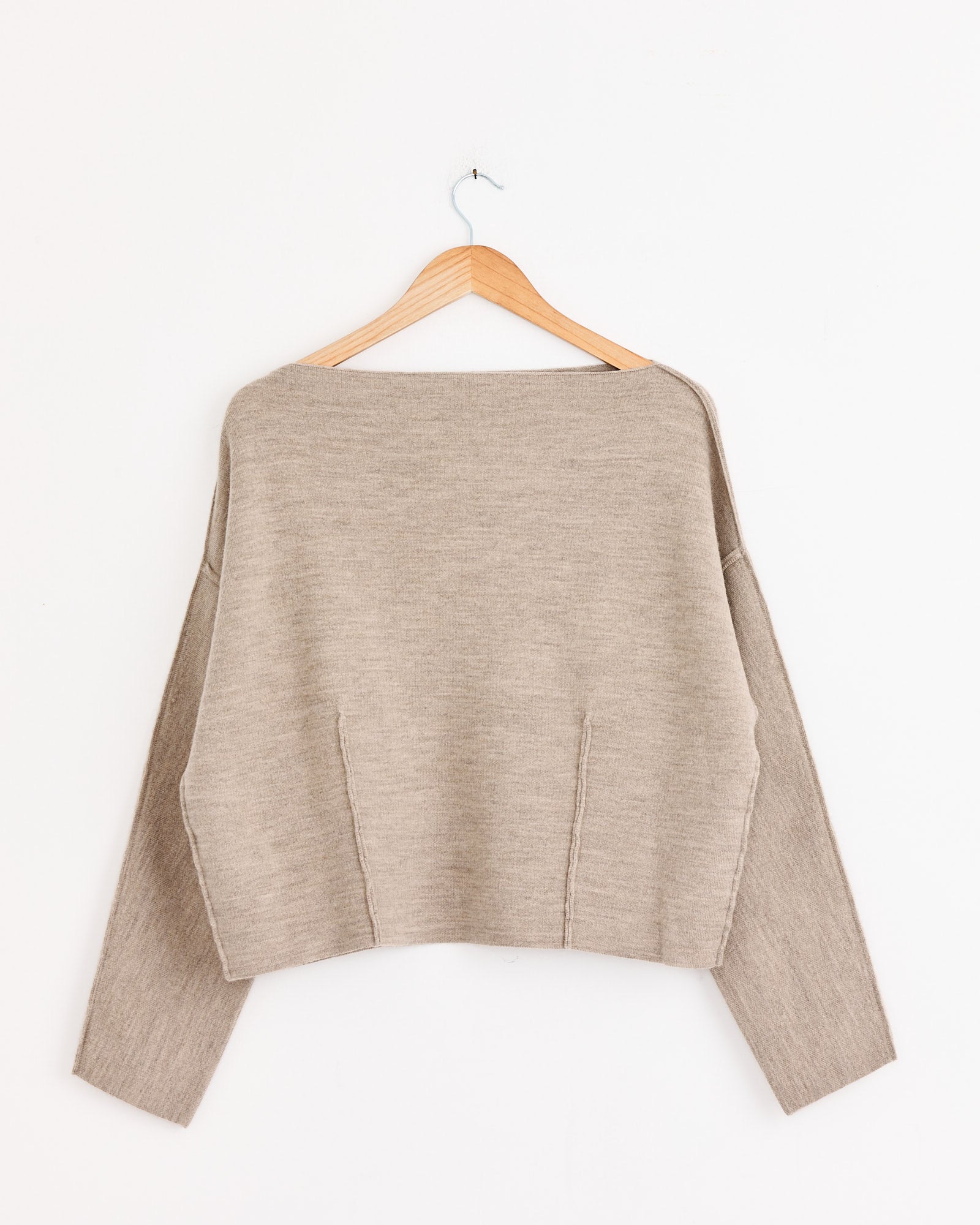 Lauren Manoogian’s Double Knit Boatneck in Oak is a beige, long-sleeve merino-alpaca sweater with a wide neckline and boxy fit, featuring simple, sculptural knitwear design. Shown on a wooden hanger against a plain white wall.
