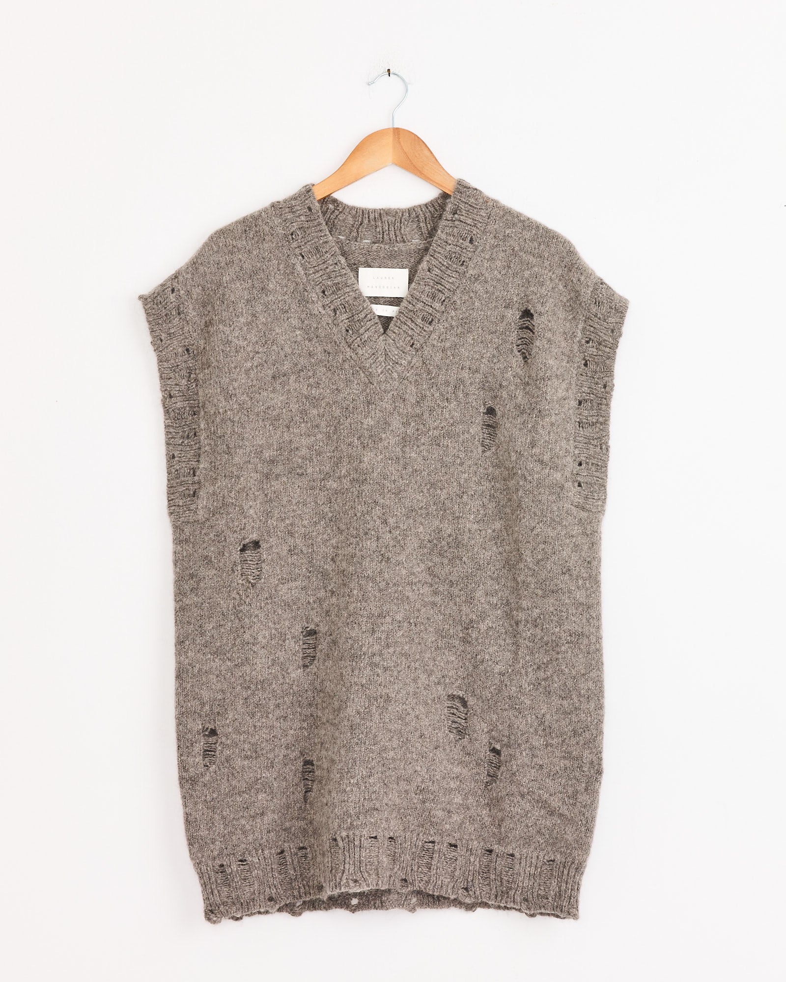A sleeveless, oversized Distressed Vest in Rock by Lauren Manoogian, with a V-neck and ripped details, hangs on a wooden hanger against a plain white wall.