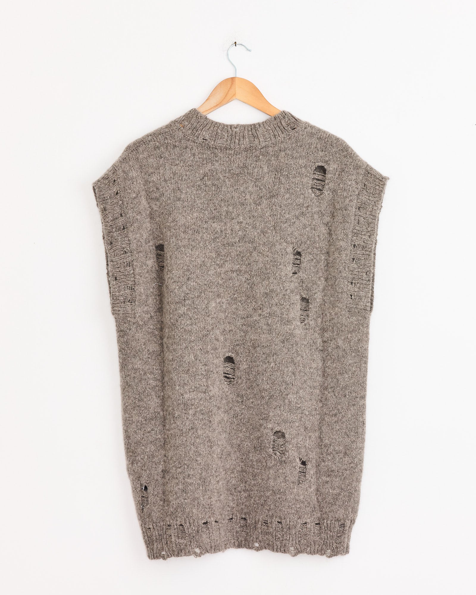 A gray, sleeveless Distressed Vest in Rock by Lauren Manoogian hangs on a wooden hanger against a white wall. Made of Merino wool, this loose-fit vest features intentional rips and distressed holes throughout the fabric.