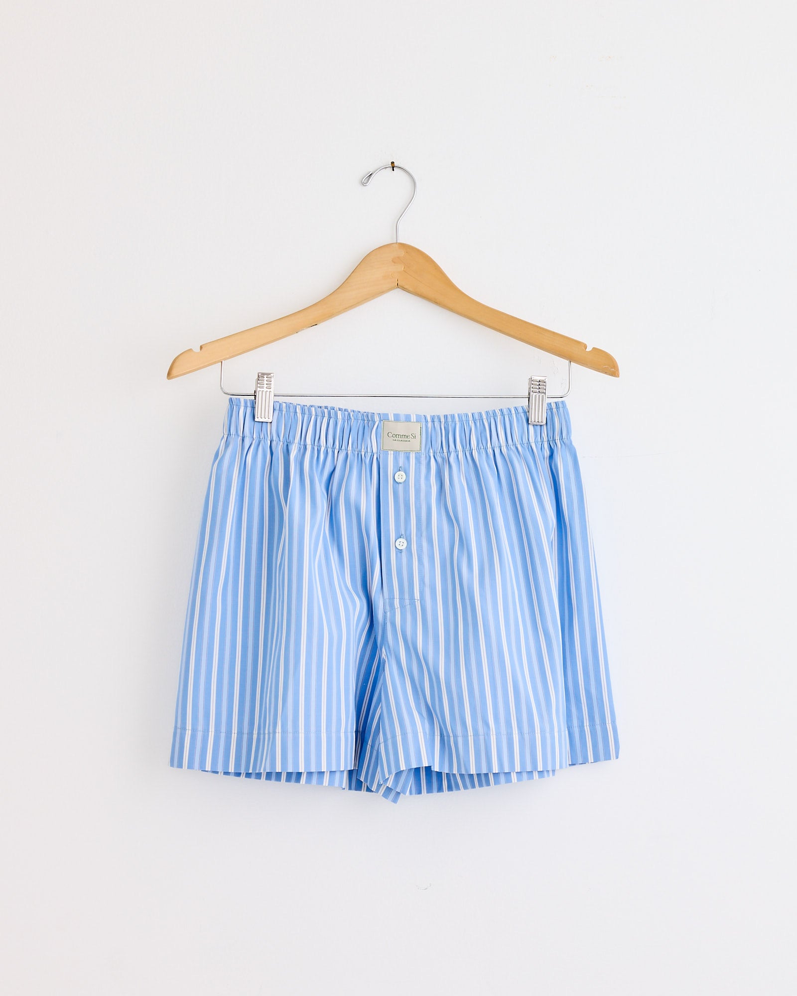 The Comme Si La Boxer Classica in Executive Stripe—light blue and white vertical stripes—hangs on a wooden hanger against a white wall. Need sizing help? Contact us via online chat for assistance.