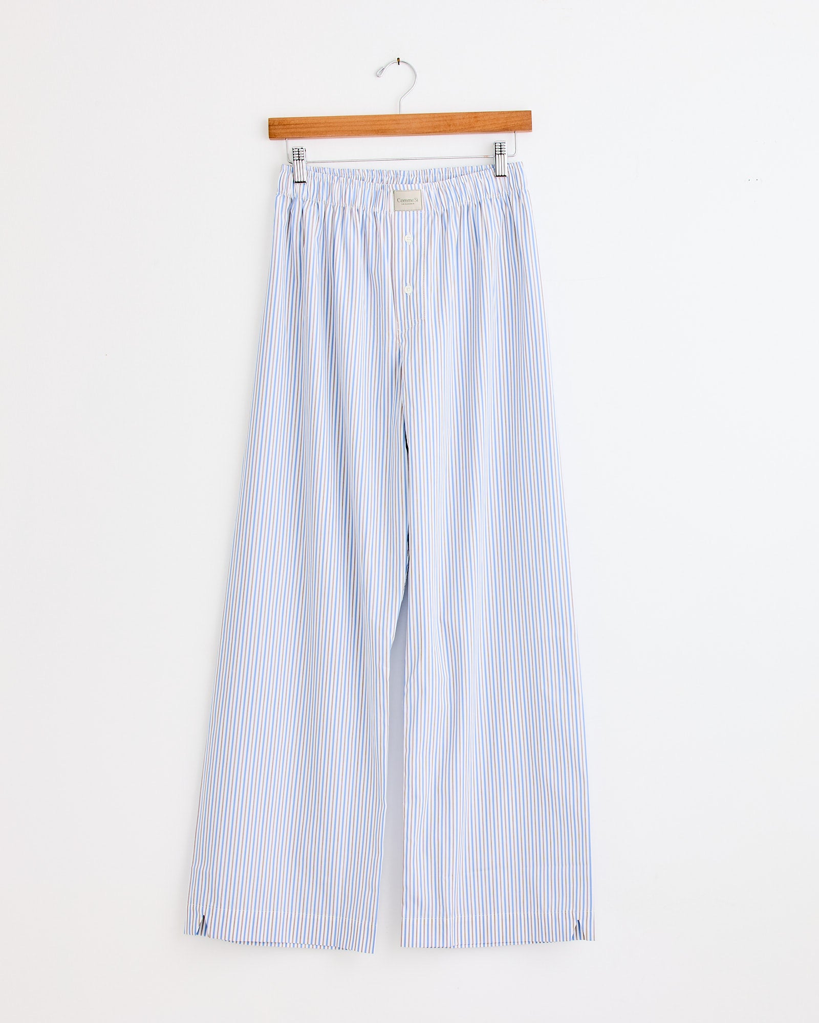The La Boxer Alta in Ledger Stripe by Comme Si features light blue and white vertical stripes, a wide-leg fit, elastic waistband, and drawstring. Hanging on a wooden hanger against a white wall. For sizing help, contact our online chat.