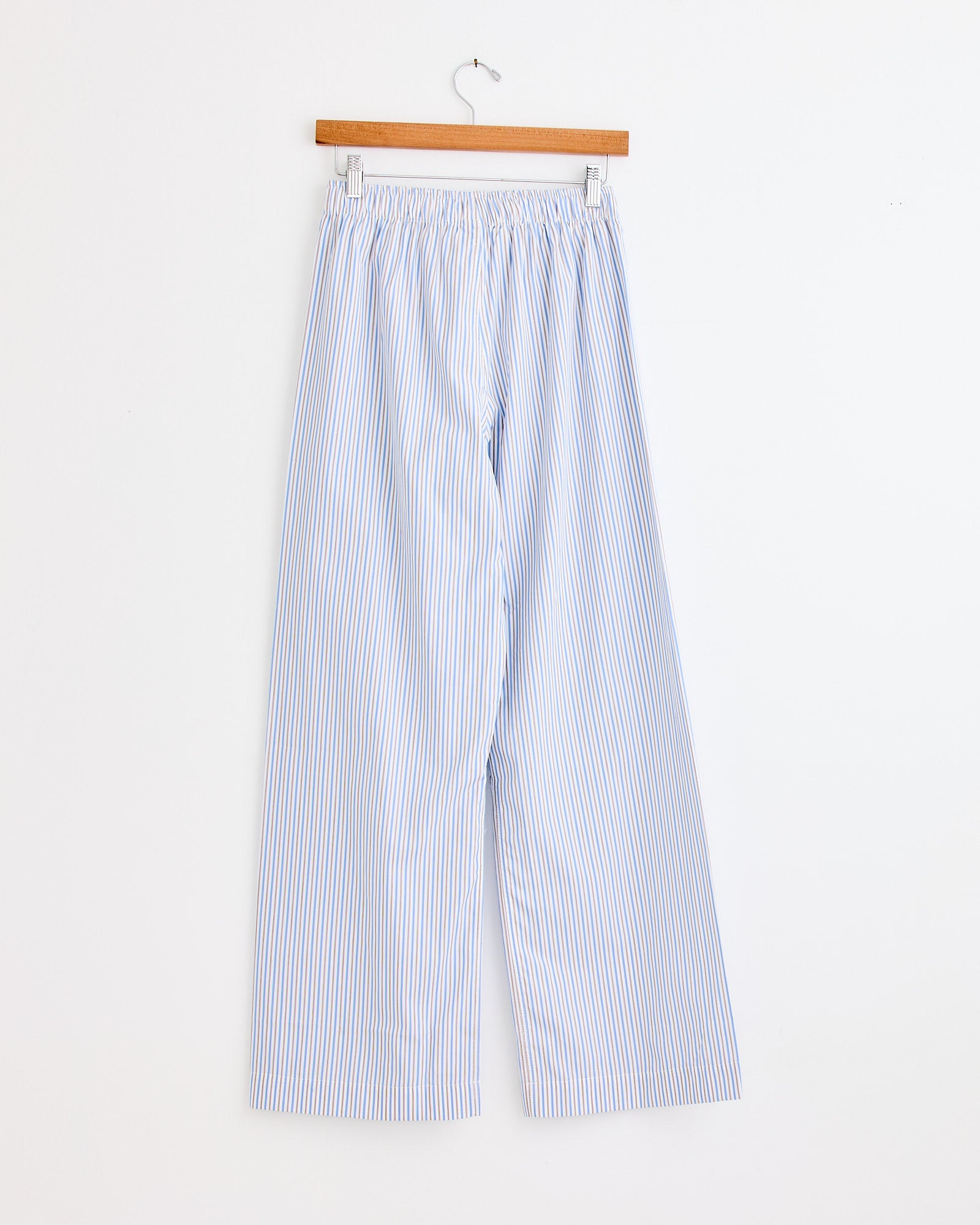 The La Boxer Alta in Ledger Stripe by Comme Si features light blue and white vertical stripes and an elastic waistband, shown hanging on a wooden hanger. For sizing or fit questions, please use our online chat.