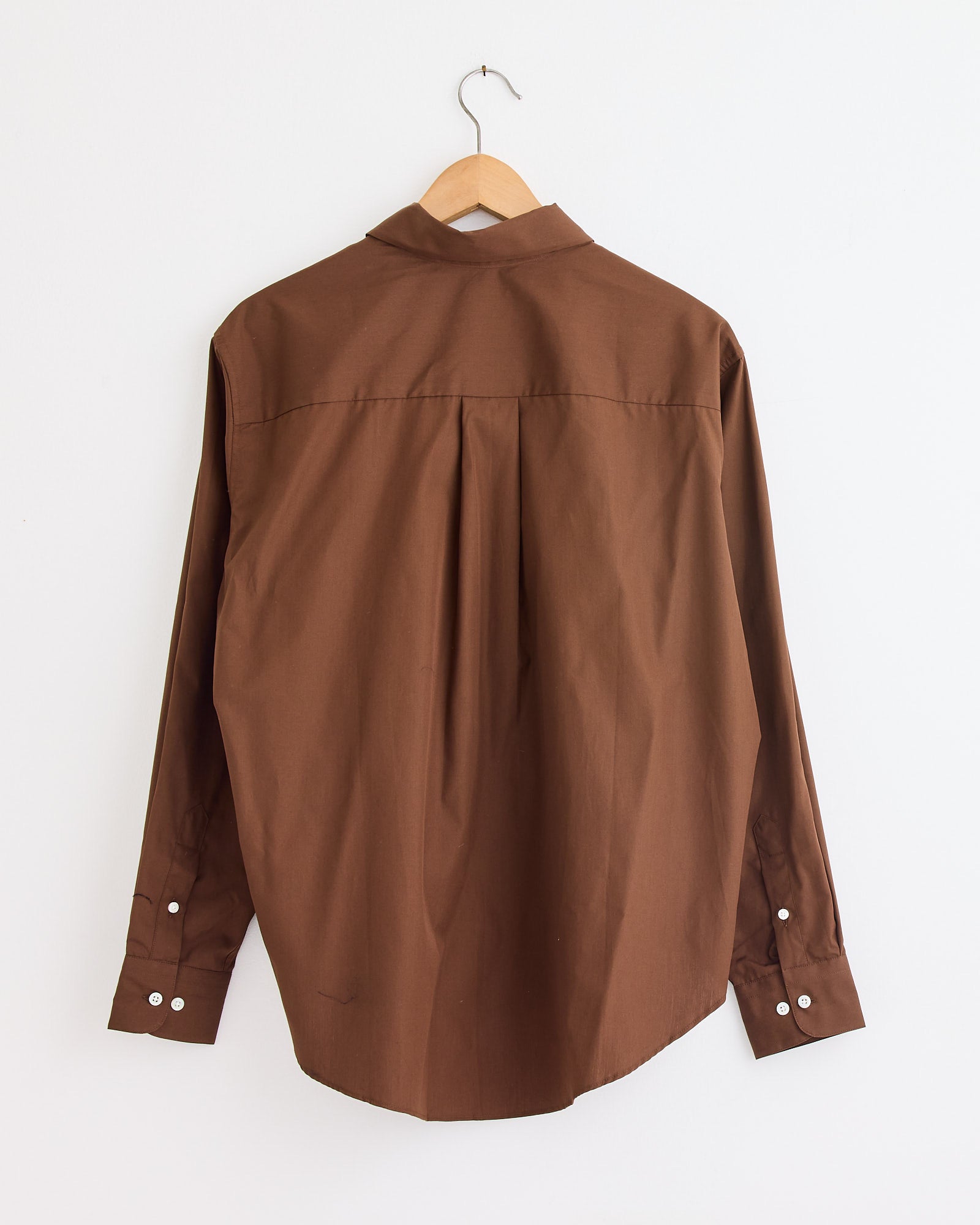The La Shirt Classica in Brown by Comme Si is shown from the back on a wooden hanger against a white wall, featuring a straight yoke, back pleat, and buttoned cuffs. Need sizing or fit help? Chat with us online.