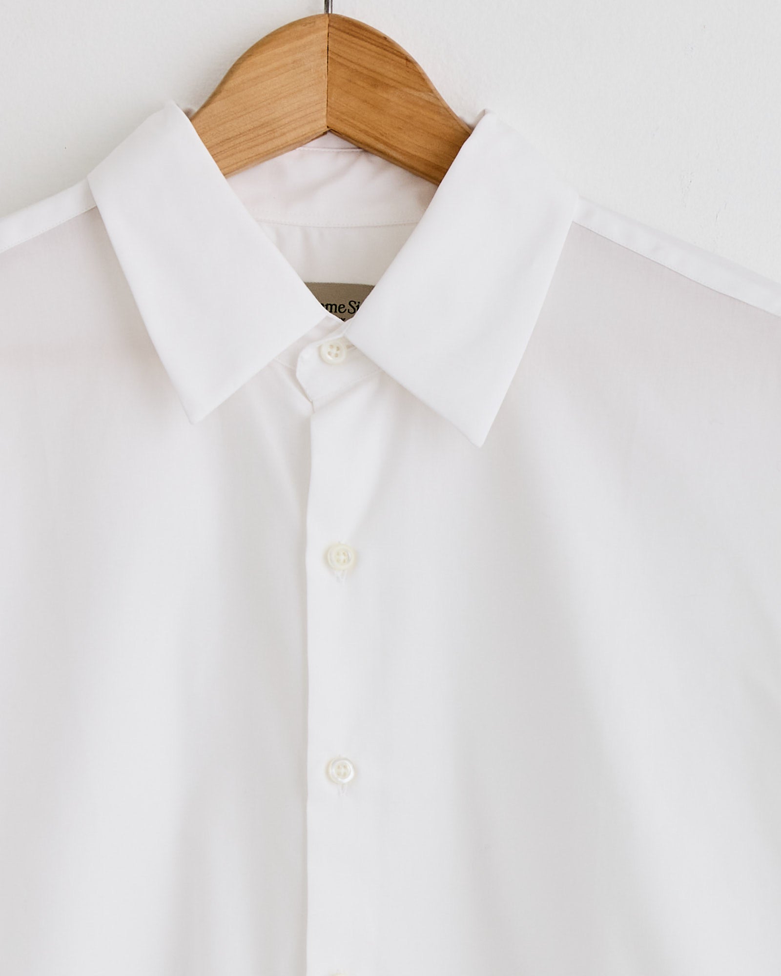 The La Shirt Classica in White by Comme Si, featuring a pointed collar, hangs neatly pressed on a wooden hanger against a plain white background—ideal for those seeking the perfect fit.
