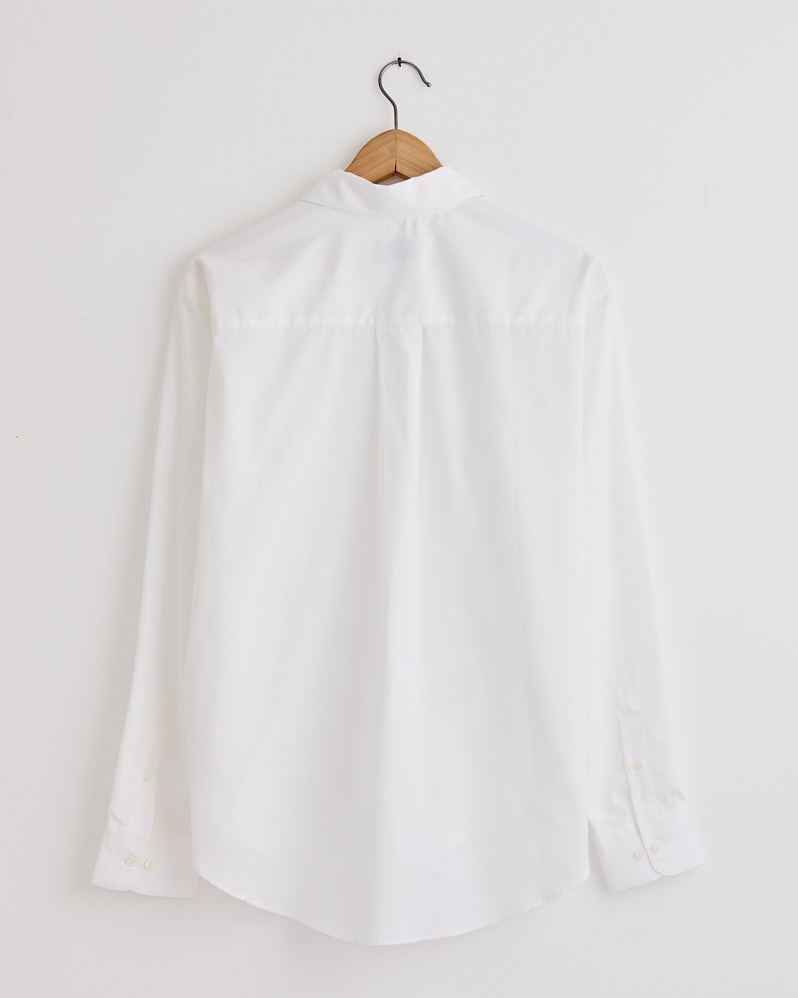 The La Shirt Classica in White by Comme Si, shown from the back on a wooden hanger against a white wall, offers timeless style and an effortless fit. For personalized sizing advice, contact our online chat.