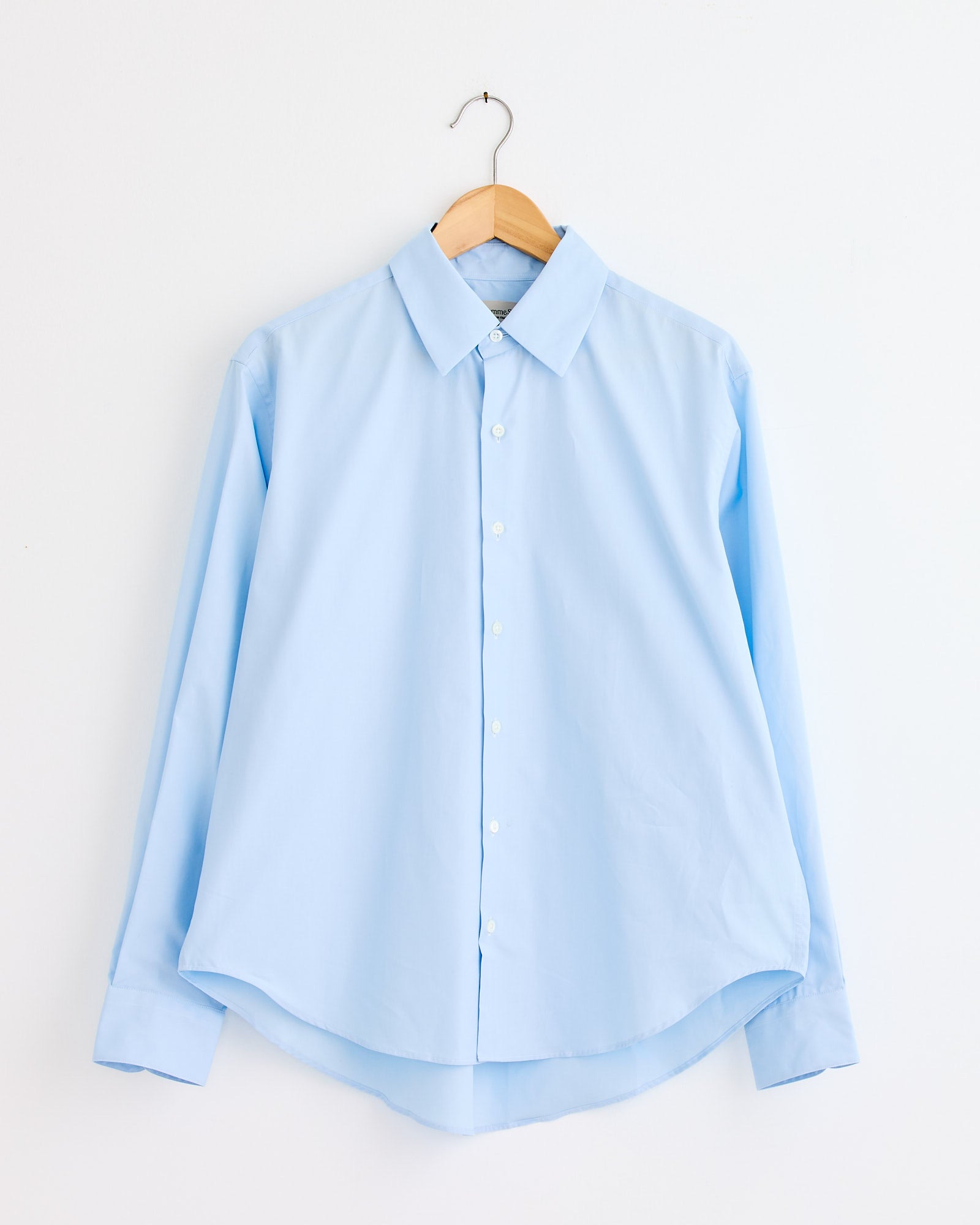 The La Shirt Classica in Marina by Comme Si—a light blue, long-sleeve button-up—hangs on a wooden hanger against a white wall. For fit or sizing questions, our online chat can help you find the perfect match.