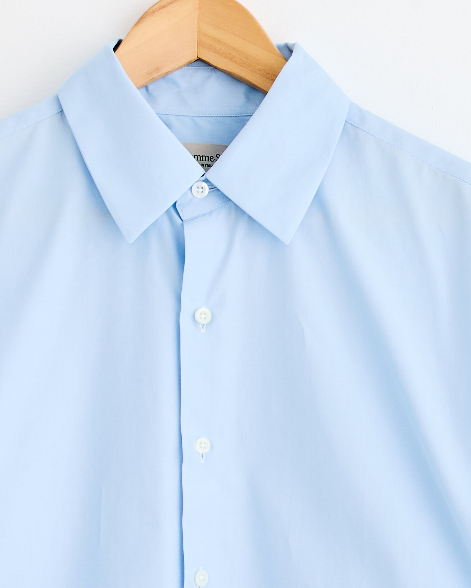 The La Shirt Classica in Marina by Comme Si—a light blue button-up with a pointed collar—hangs on a wooden hanger against a white background, ideal for achieving the perfect fit.