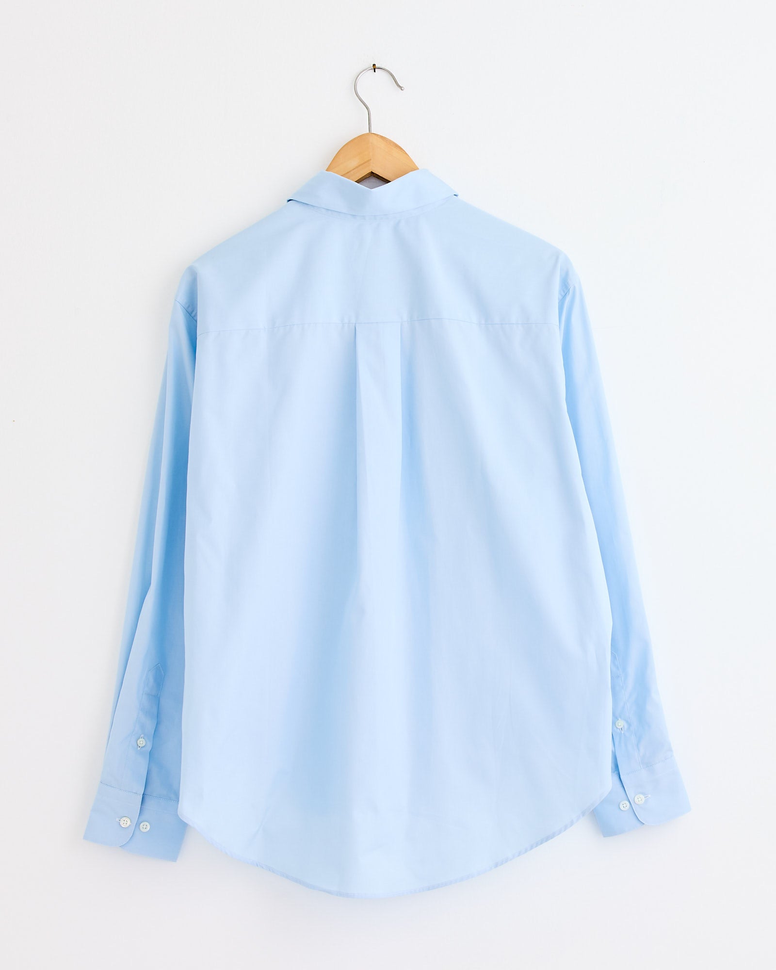 The Comme Si La Shirt Classica in Marina, a light blue long-sleeve dress shirt, hangs on a wooden hanger against a white wall (back view). For fit or sizing questions, our online chat is here to help.