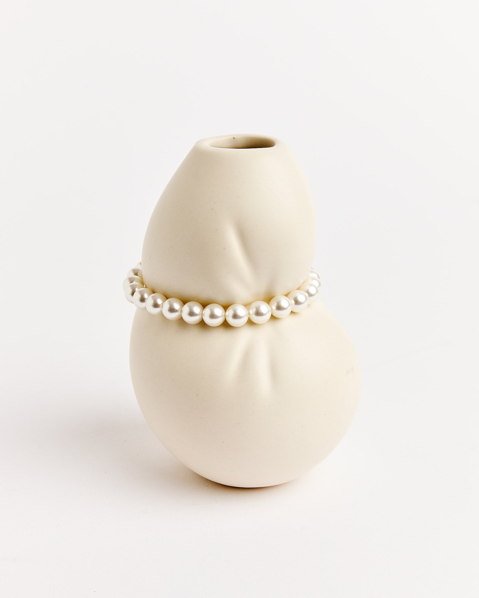 The Small Vessel With Faux Pearls by Completedworks is a smooth, off-white, abstract vase featuring a single strand of faux pearls around its center, set against a plain white background.