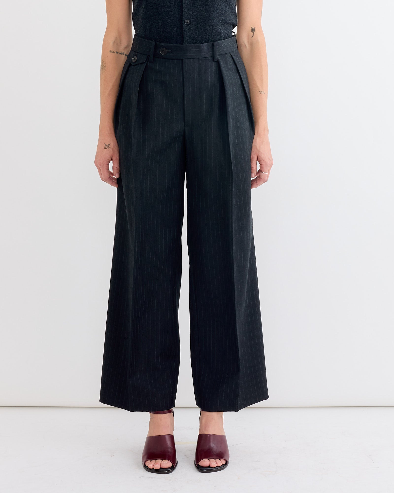 Only the lower torso and legs are visible as a person stands against a plain white background, wearing Auralee Bluefaced Two Tuck Slack in Charcoal Navy Stripe paired with burgundy open-toe heels.