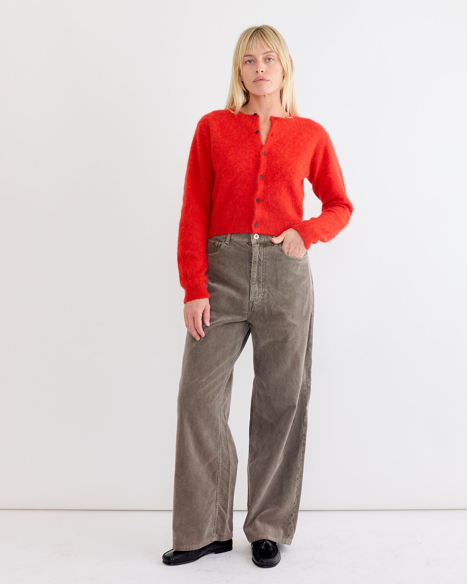 A woman with blonde hair and bangs stands against a white background, wearing the Auralee Baby Alpaca Cardigan in Red, loose taupe pants, and black shoes. She looks slightly to the side with one hand in her pocket.