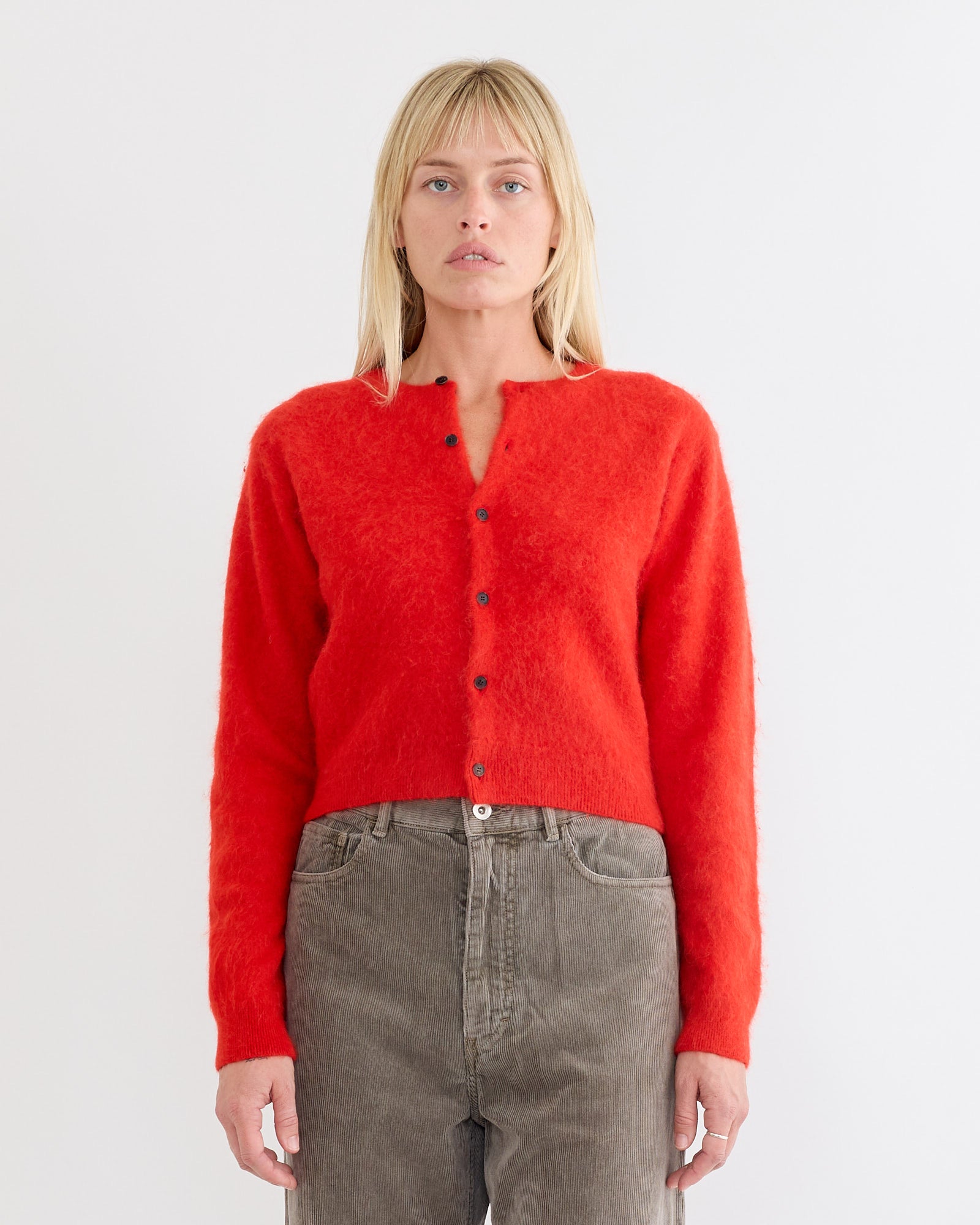 A woman with blonde hair wears the Auralee Baby Alpaca Cardigan in Red with high-waisted gray jeans, posing against a white background and looking directly at the camera.