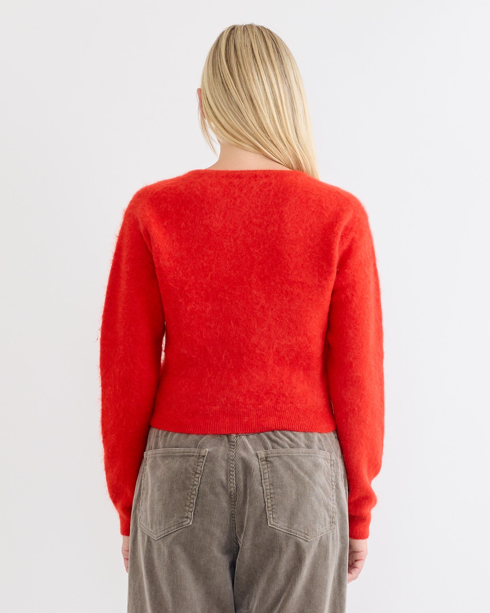 A person with long blonde hair, seen from behind, wears the Auralee Baby Alpaca Cardigan in red with loose-fitting gray corduroy pants against a plain white background.