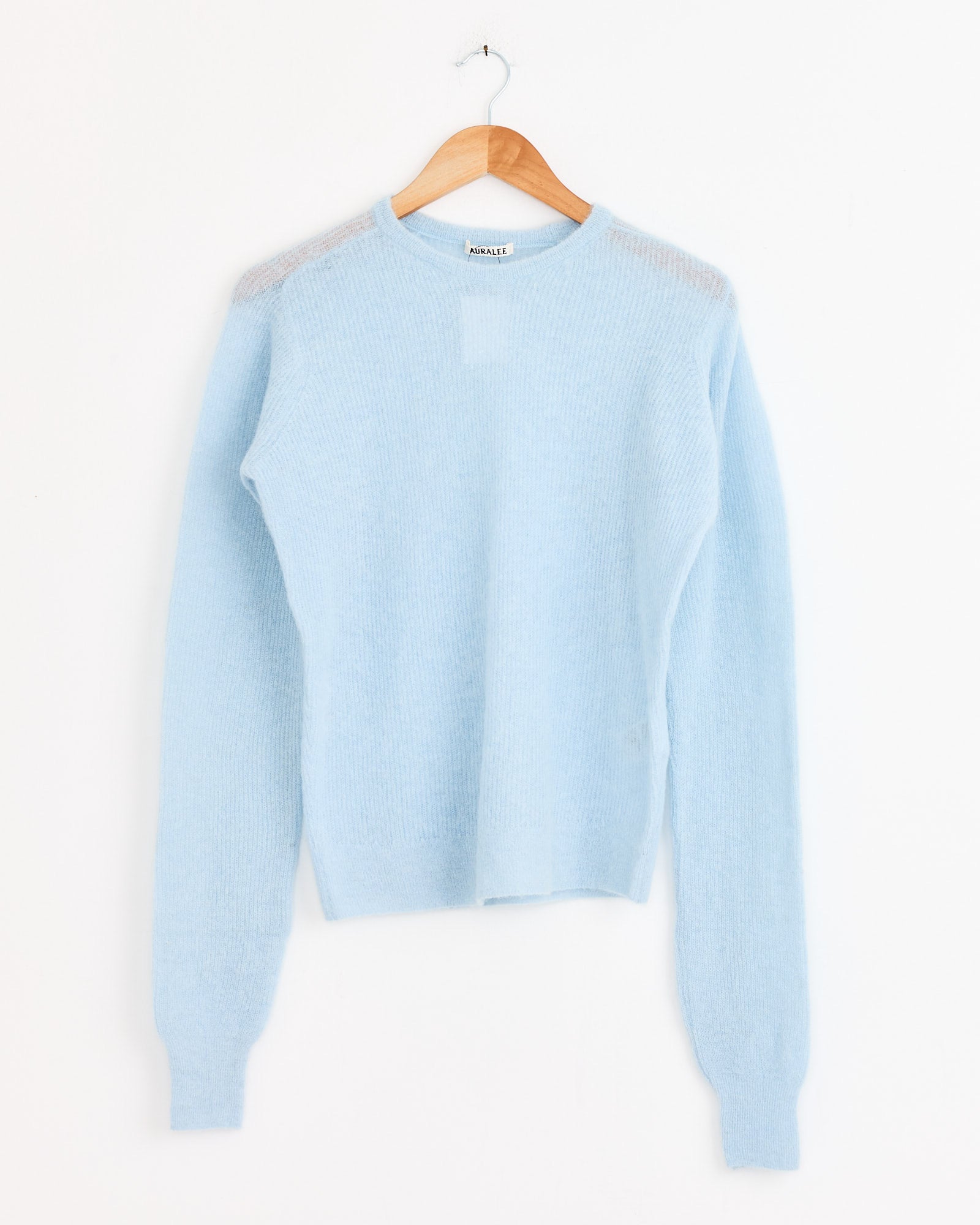 The Auralee Baby Suri Alpaca Pullover in Light Blue, a luxurious long-sleeve knit sweater, is displayed on a wooden hanger against a plain white background.