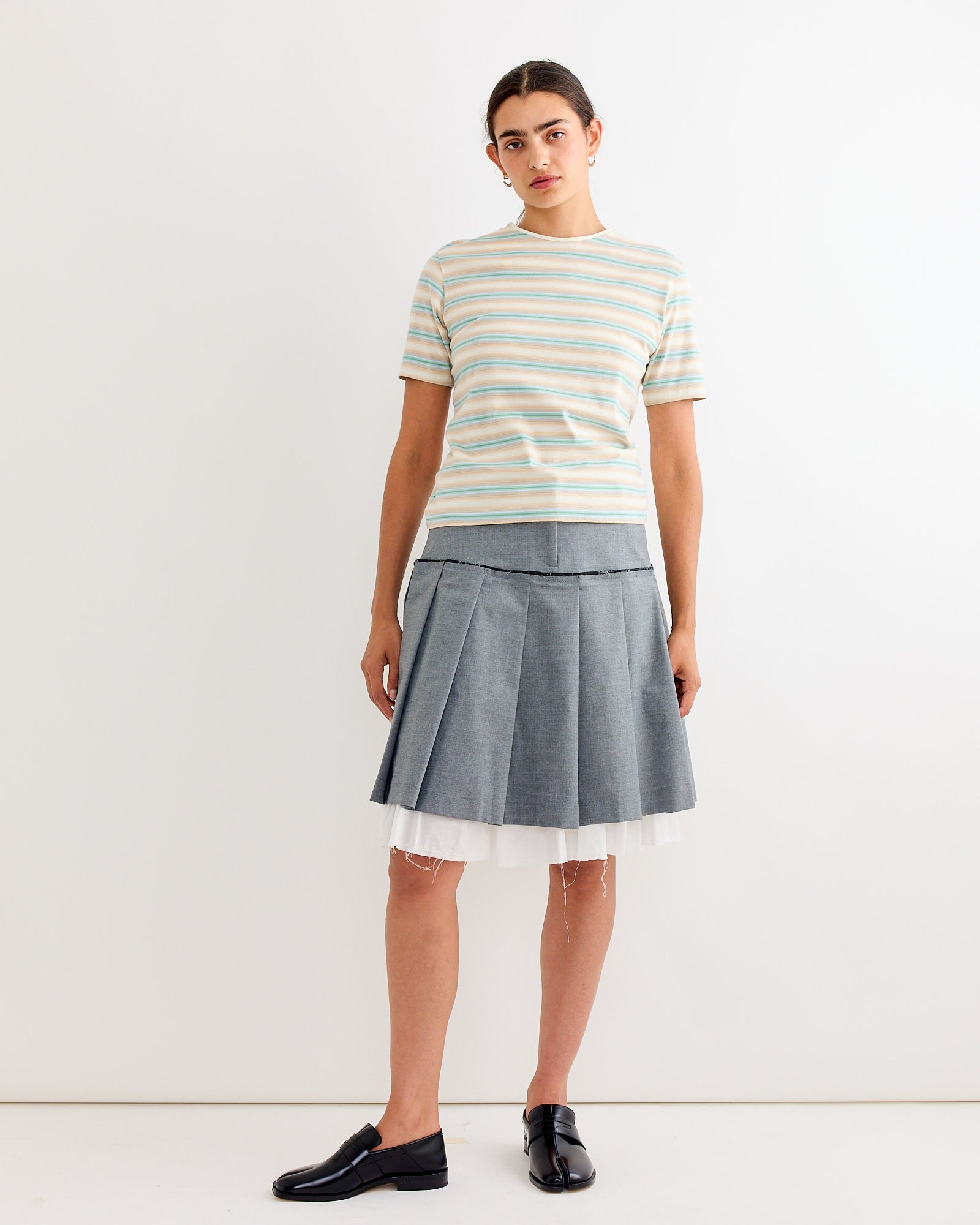 A woman wearing Auralees Gradient Jersey Tee in Fade Blue Stripe, a gray pleated skirt with white tulle, and black loafers stands against a plain white background, looking straight ahead with a neutral expression.
