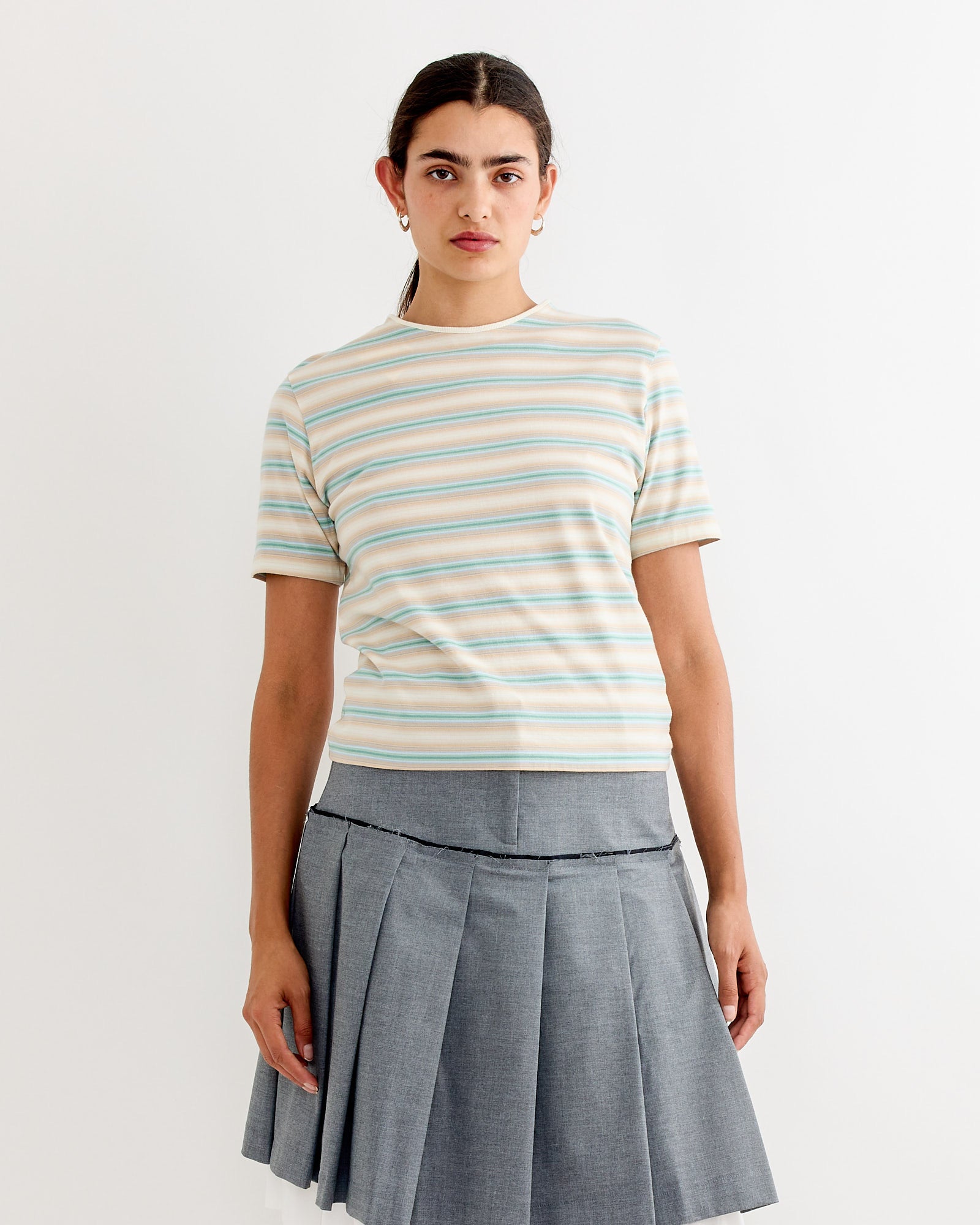 A person stands against a white background wearing Auralee’s Gradient Jersey Tee in Fade Blue Stripe and a gray pleated skirt. Their hair is pulled back, with a neutral expression. The cotton jersey fabric creates a laid-back look.