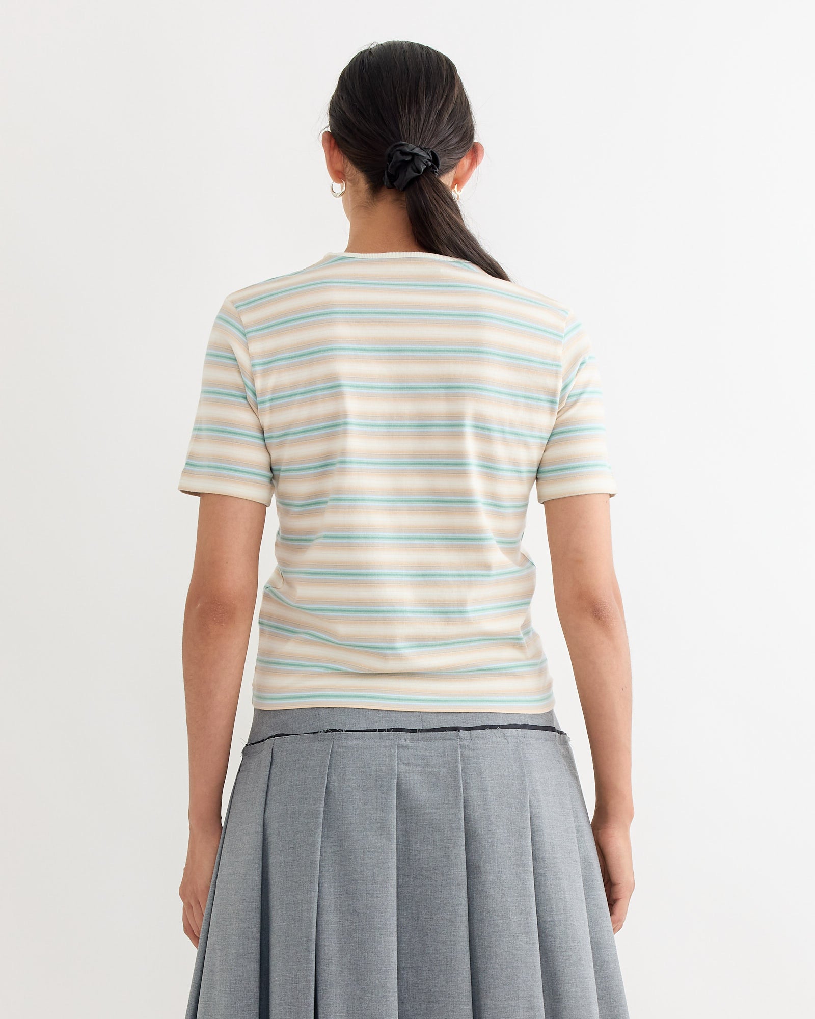 A person with long dark hair in a ponytail, wearing the Auralee Gradient Jersey Tee in Fade Blue Stripe and a gray pleated skirt, stands with their back to the camera against a plain white background.