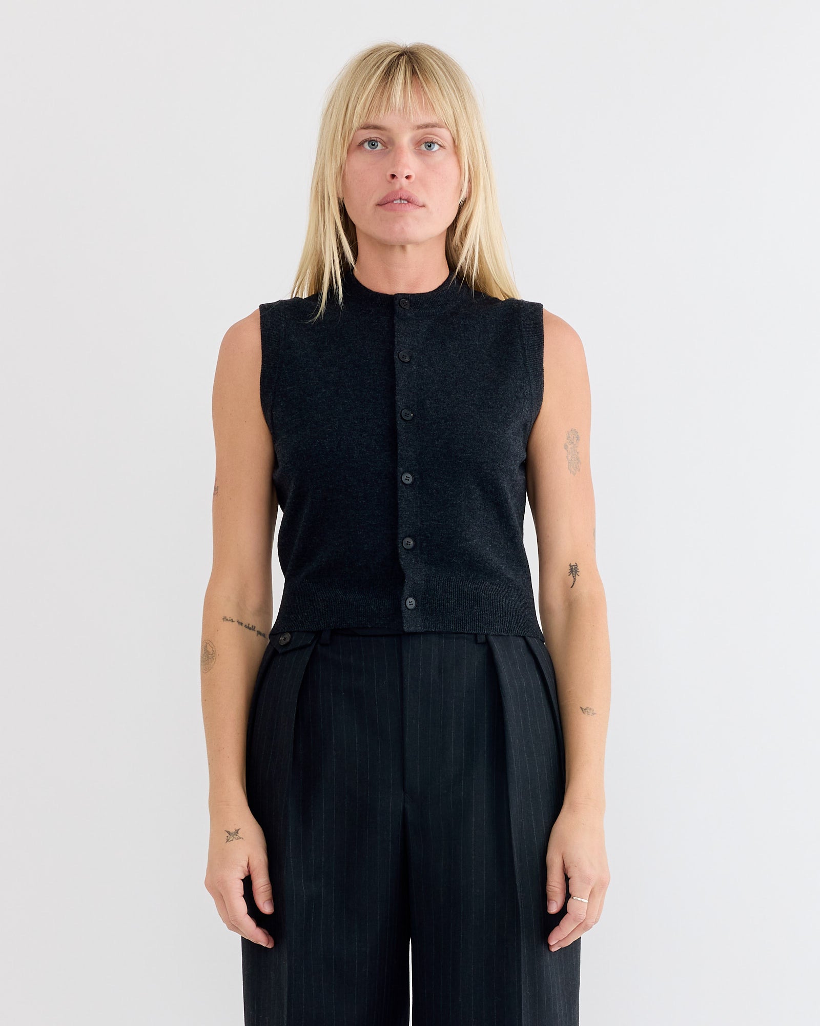 A woman with straight blonde hair and arm tattoos wears the Auralee High Twist Wool Vest in Charcoal and high-waisted black pants, standing against a plain white background and looking directly at the camera.