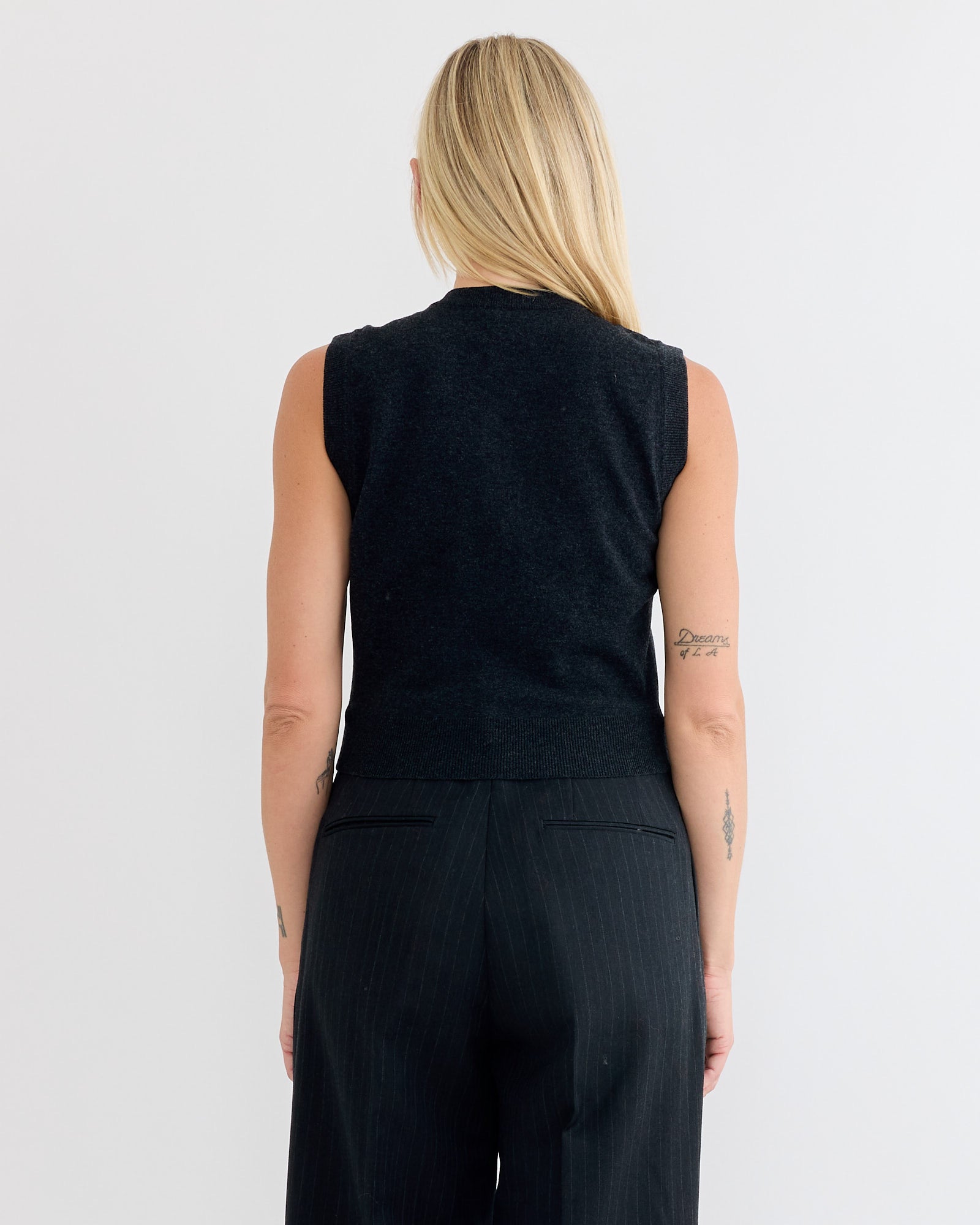 A person with long blonde hair, seen from behind, wears the Auralee High Twist Wool Vest in Charcoal and black pinstripe pants. Small arm tattoos are visible. The background is plain white.