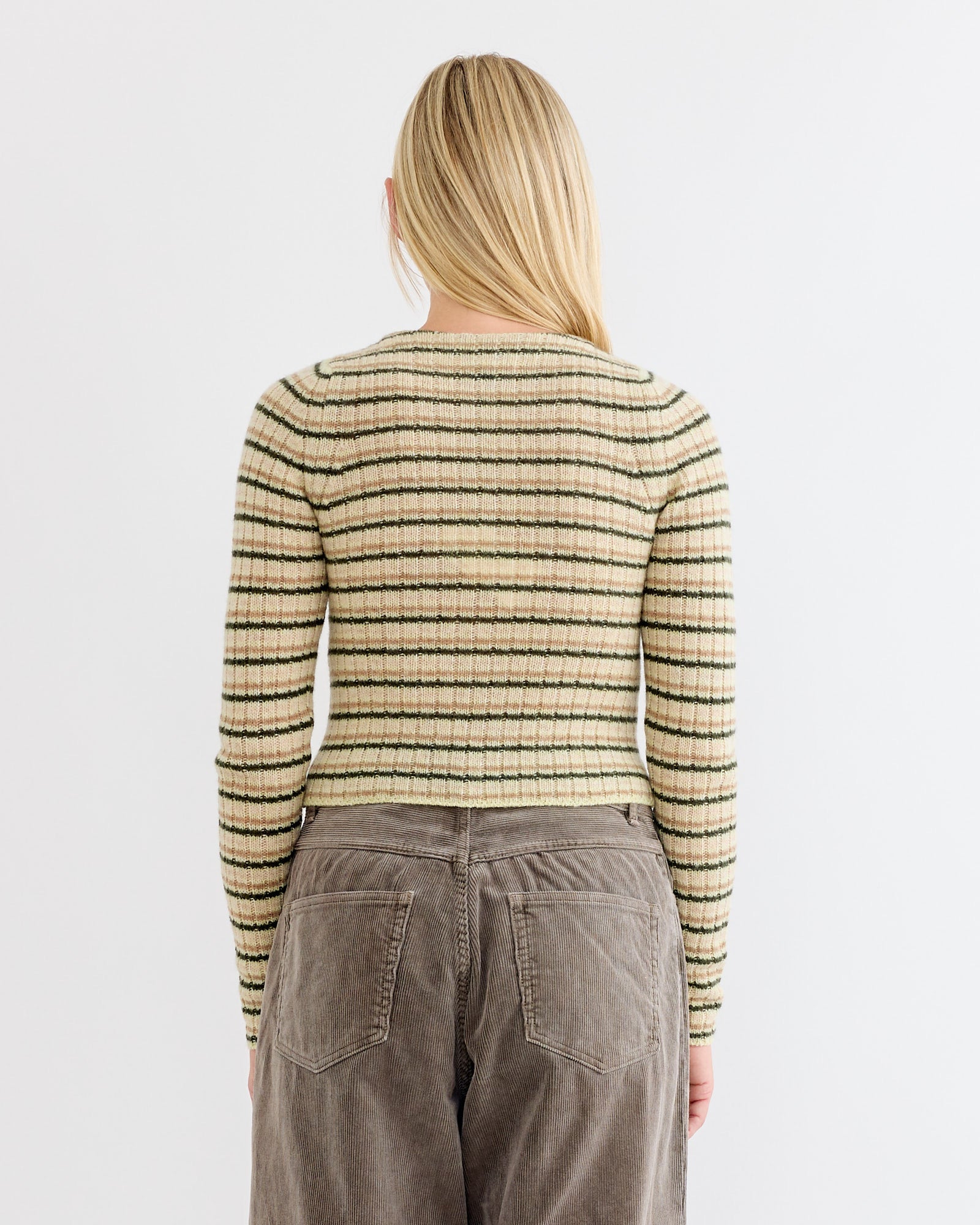 A person with long blonde hair is shown from the back, wearing the Auralee Wool Cashmere Boat Neck Pullover in Beige Stripe and high-waisted brown corduroy pants. The background is plain white. Made in Japan.
