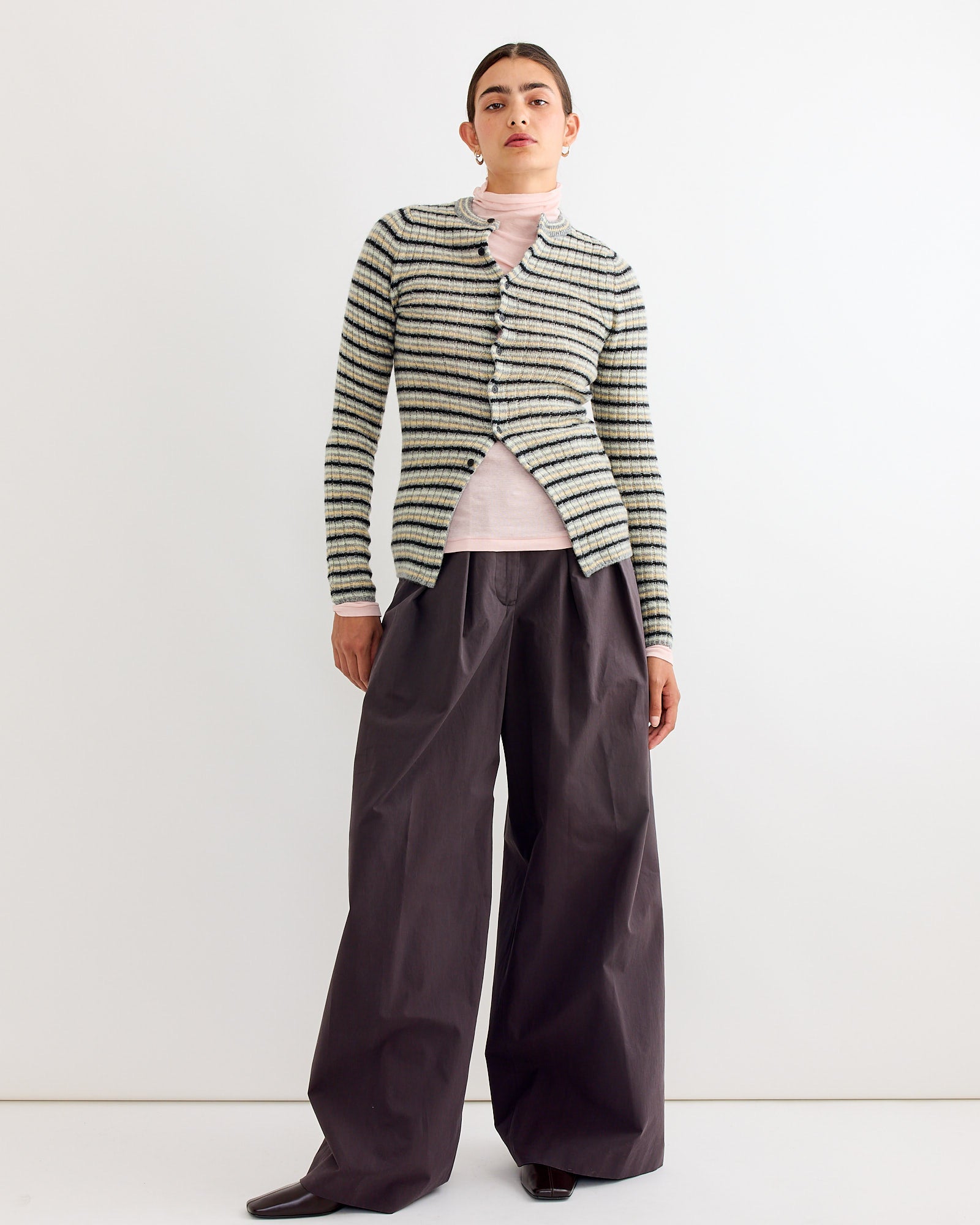 A person stands against a plain white background, wearing the Auralee Wool Cashmere Cardigan in Grey Stripe over a light pink turtleneck, paired with wide-legged dark gray pants and black shoes.