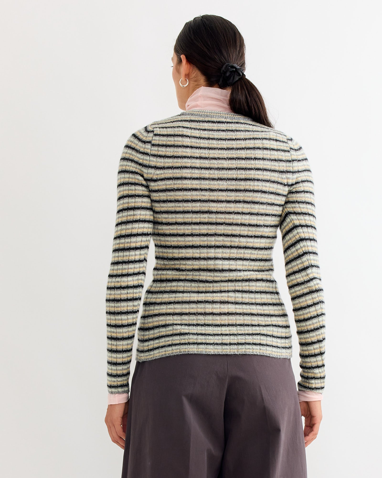 A woman with dark hair in a low ponytail faces away, wearing the Auralee Wool Cashmere Cardigan in Grey Stripe over a pale pink turtleneck and dark wide-legged pants, set against a plain white background.