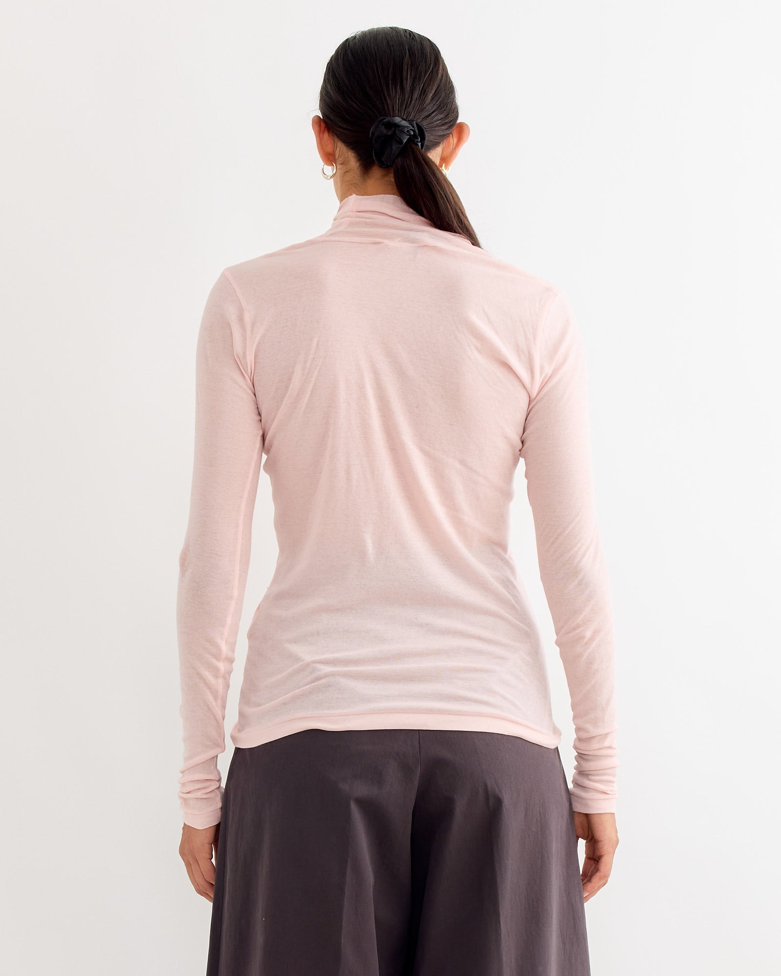 A person with long dark hair in a low ponytail is seen from behind, wearing the Auralee Sheer Rib Turtleneck Tee in Light Pink and wide-leg dark pants, standing against a plain white background.