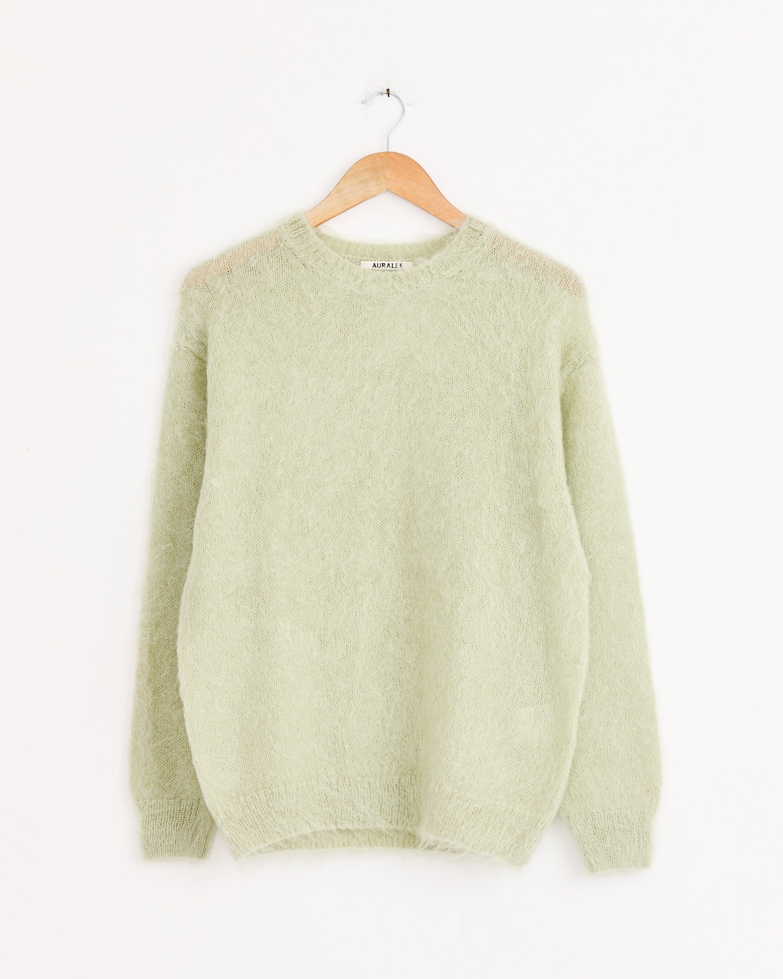 The Auralee Brushed Super Kid Mohair Pullover in Light Khaki, featuring long sleeves and a fuzzy texture, hangs on a wooden hanger against a plain white background.