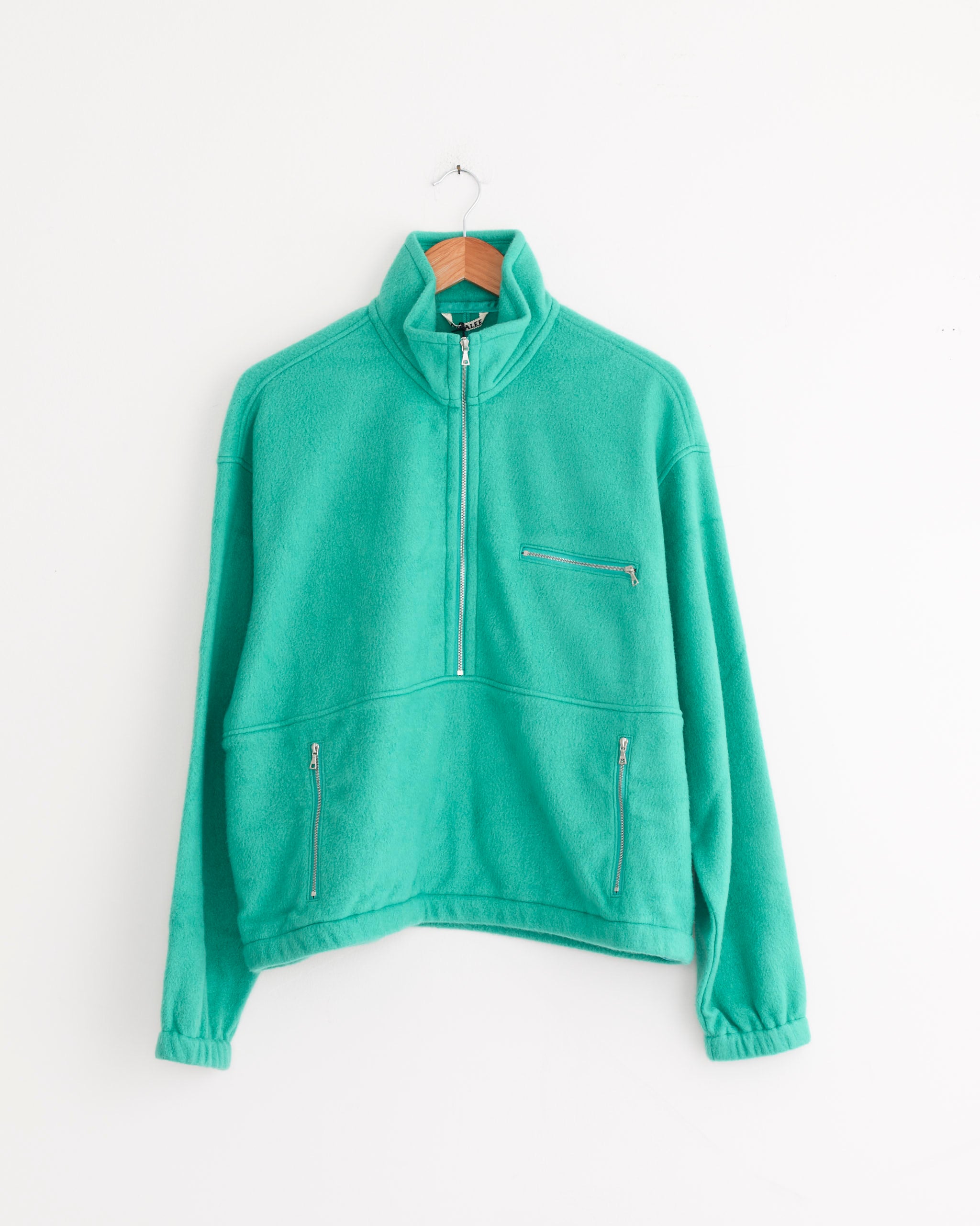 The Auralee Pure Silk Half Zip Pullover in Jade Green features a high collar, zippered chest and side pockets, and hangs on a wooden hanger against a white wall. Crafted in Japan for exceptional quality and style.